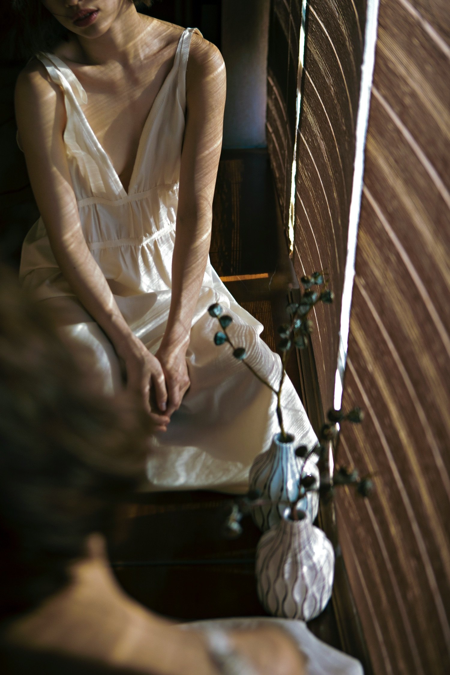 A woman sitting on the floor wearing a light-colored dress in a room with wooden walls, with shadows cast by sunlight coming through a window, and a vase with decorative branches nearby.