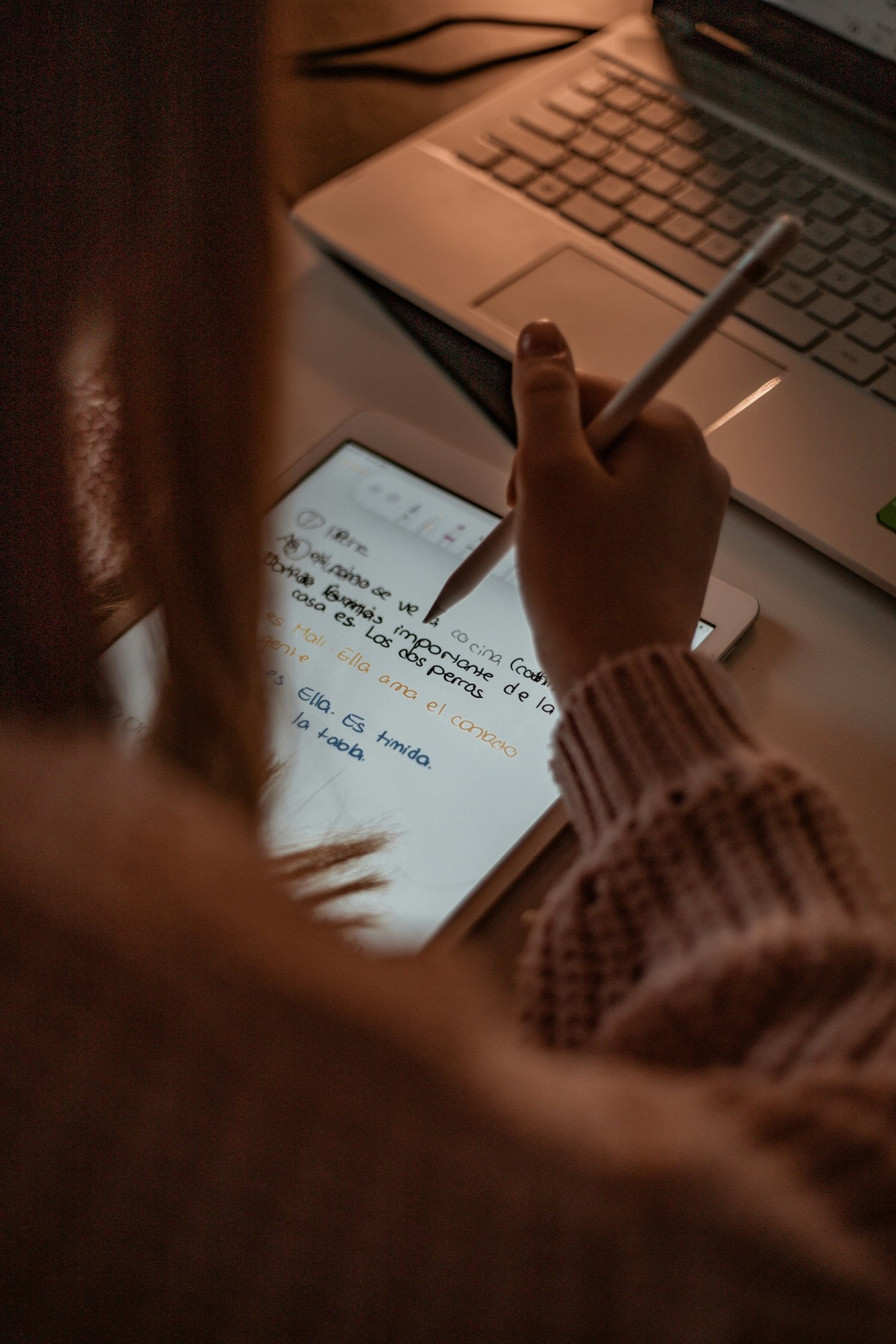 Person writing on a digital tablet with a stylus, with a laptop in the background.