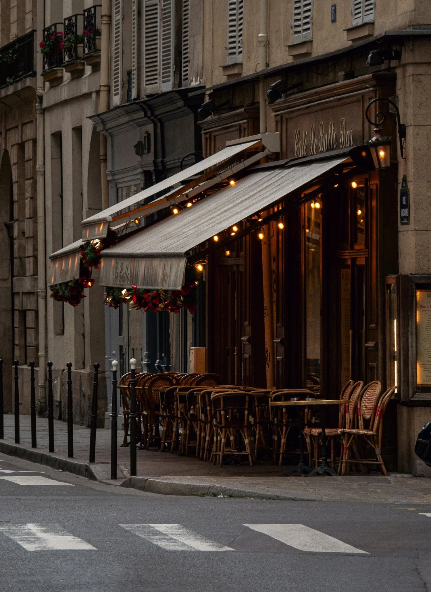 Empty outdoor seating area of a Parisian-style cafe with chairs and tables, decorated with Christmas garlands, on a street corner during evening.