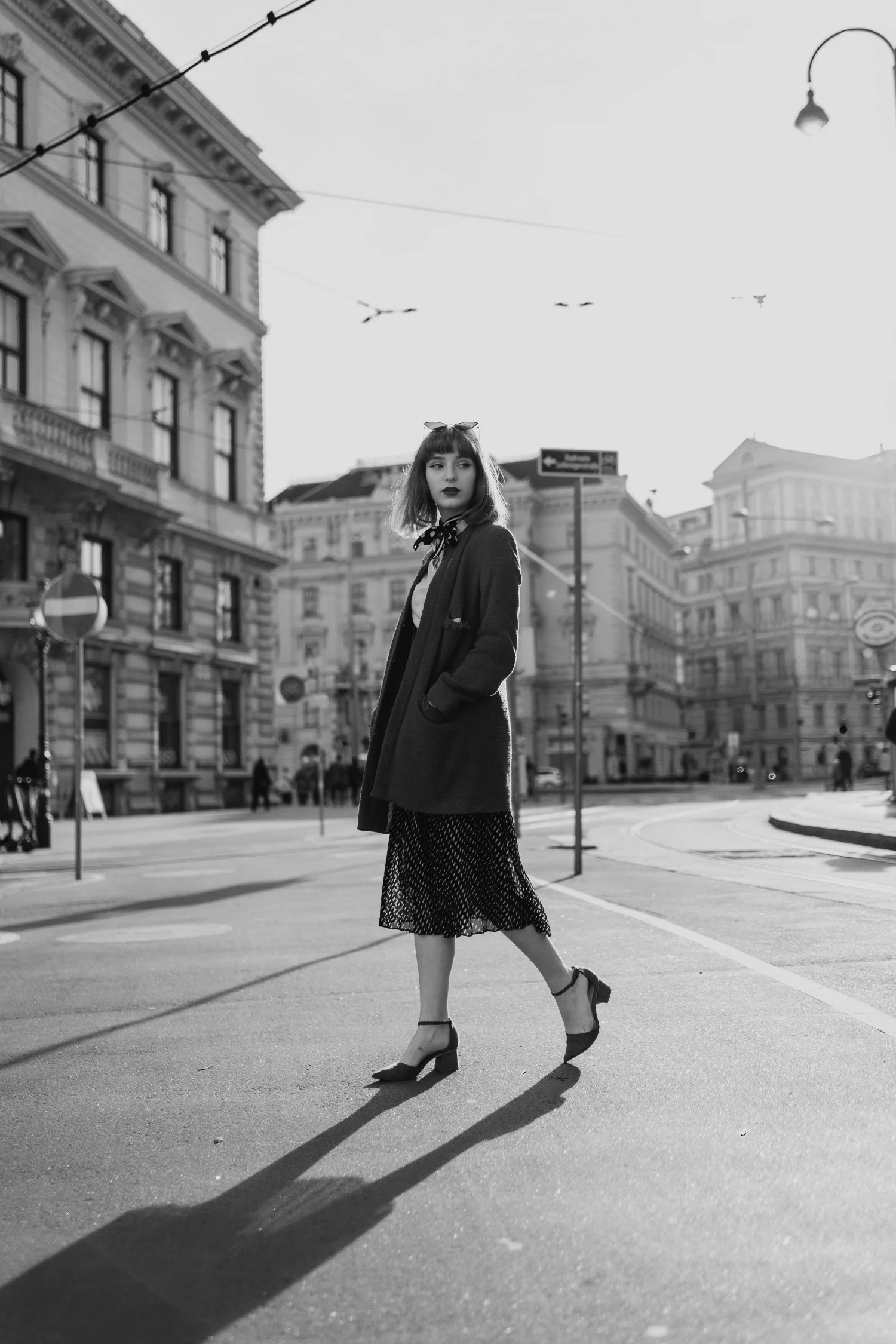 Black and white photo of a woman walking across a city street with historic buildings in the background, wearing a coat, skirt, and heels, with her shadow cast on the pavement.