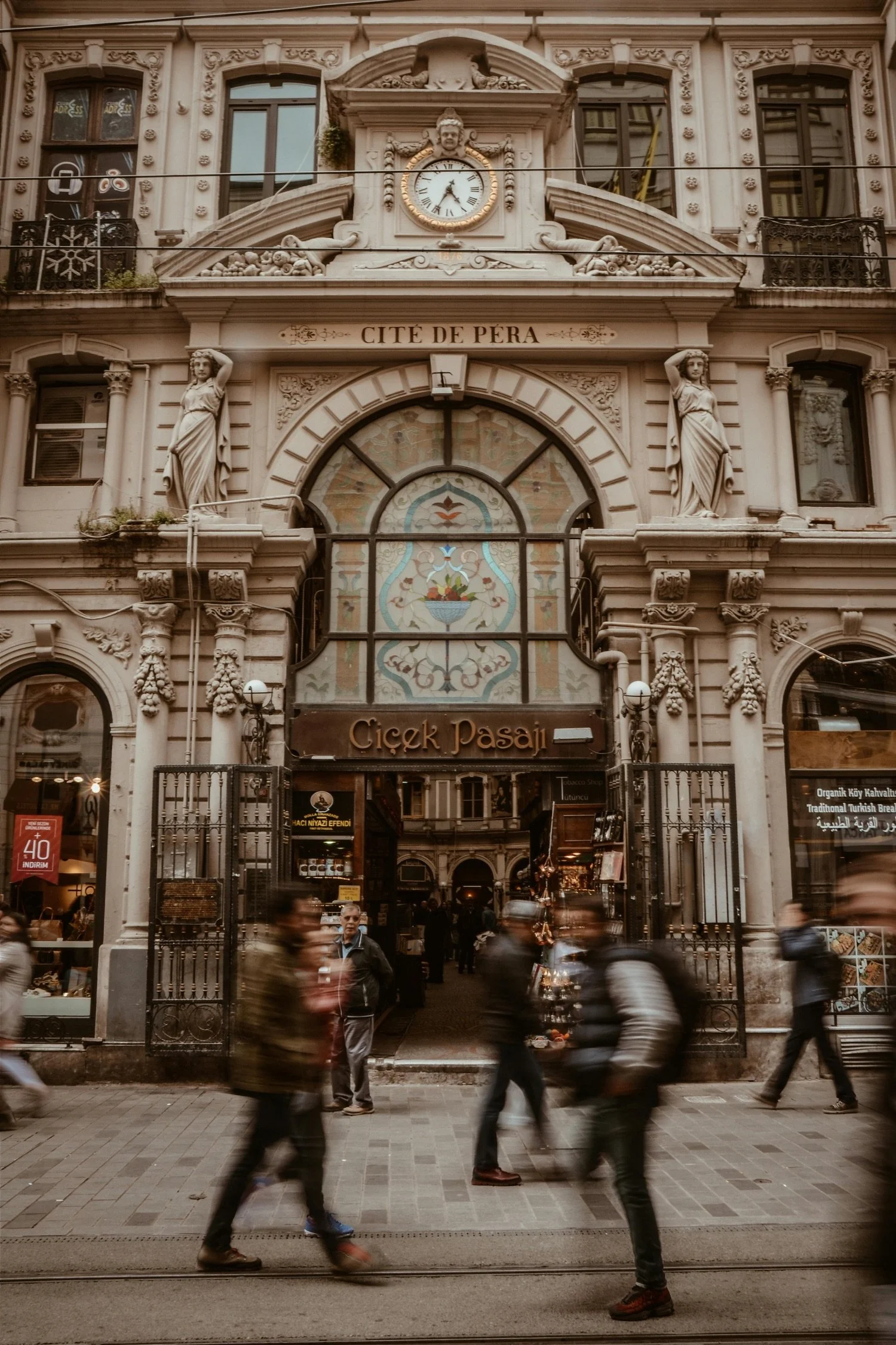 Historic building with ornate architecture, statues, clock, and stained glass entrance, sign reading 'Cité de Pera' and 'Çiçek Pasajı'; people walking in front.