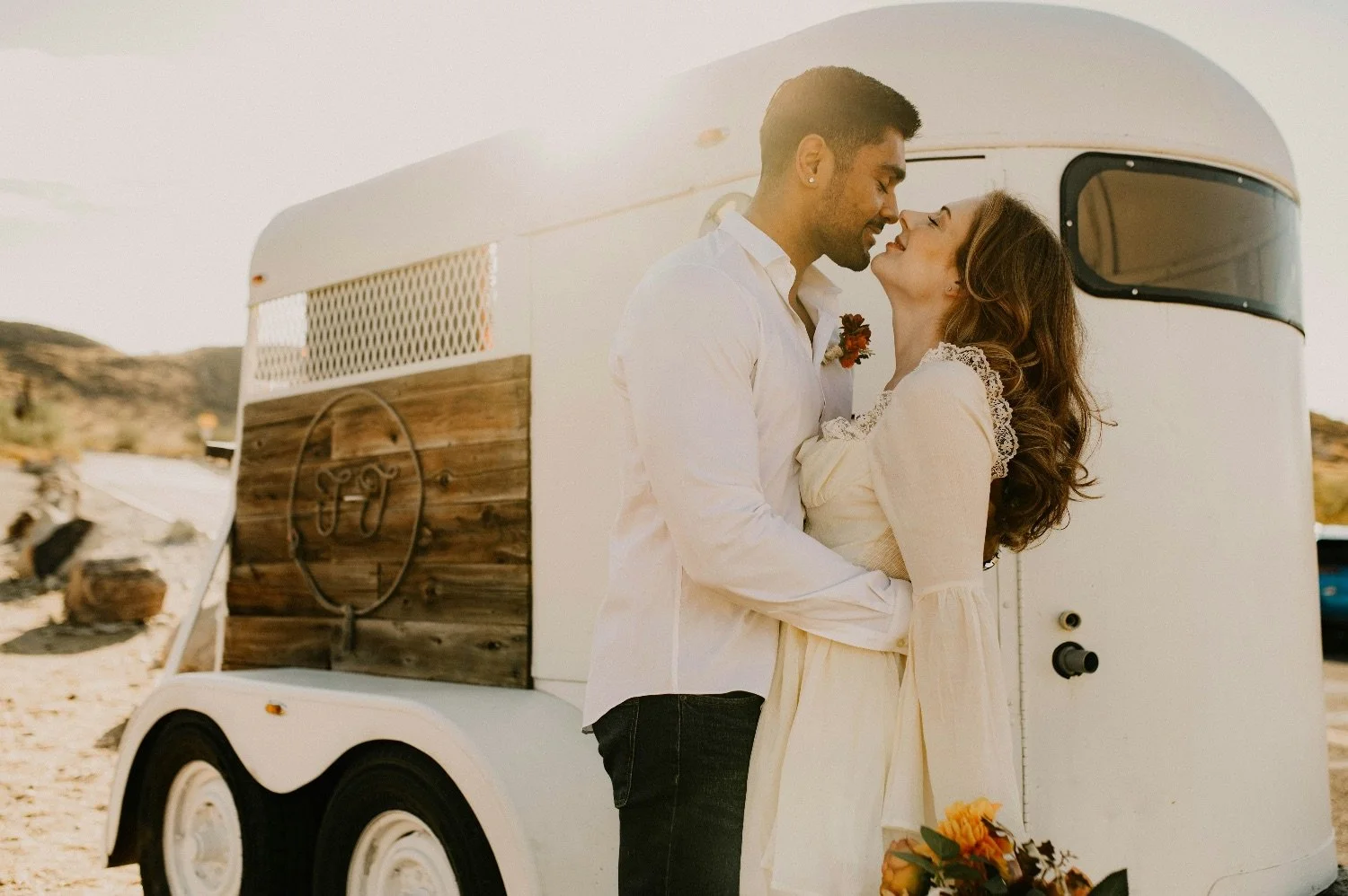 A couple embraces outdoors in front of a vintage enamel trailer, with a desert landscape in the background, during sunset.