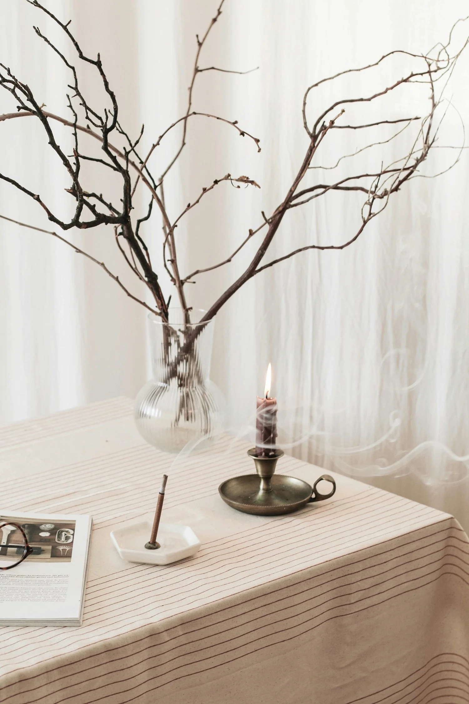 A table decorated with a striped tablecloth, a vintage brass candle holder with a lit candle, a white dish with a single stick, a pair of glasses, an open magazine, and a large glass vase with bare, twisted tree branches. White curtains are in the background.