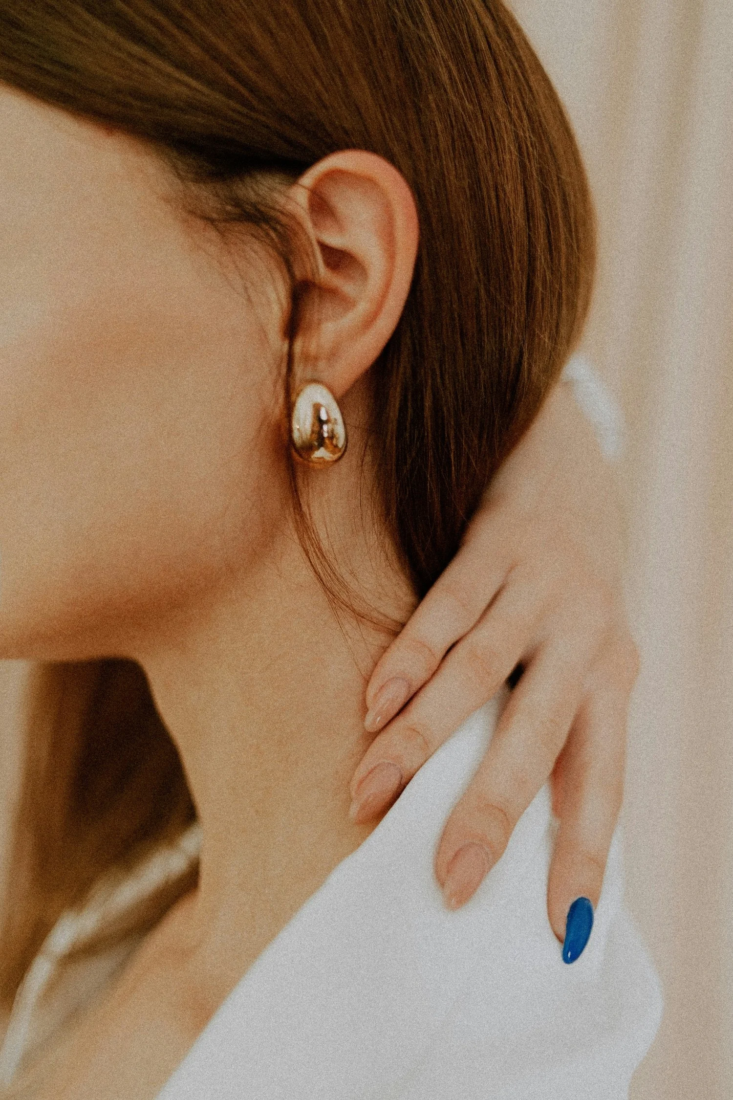 Close-up of a woman’s ear with a gold earring, her hand resting on her neck, and nails painted blue.