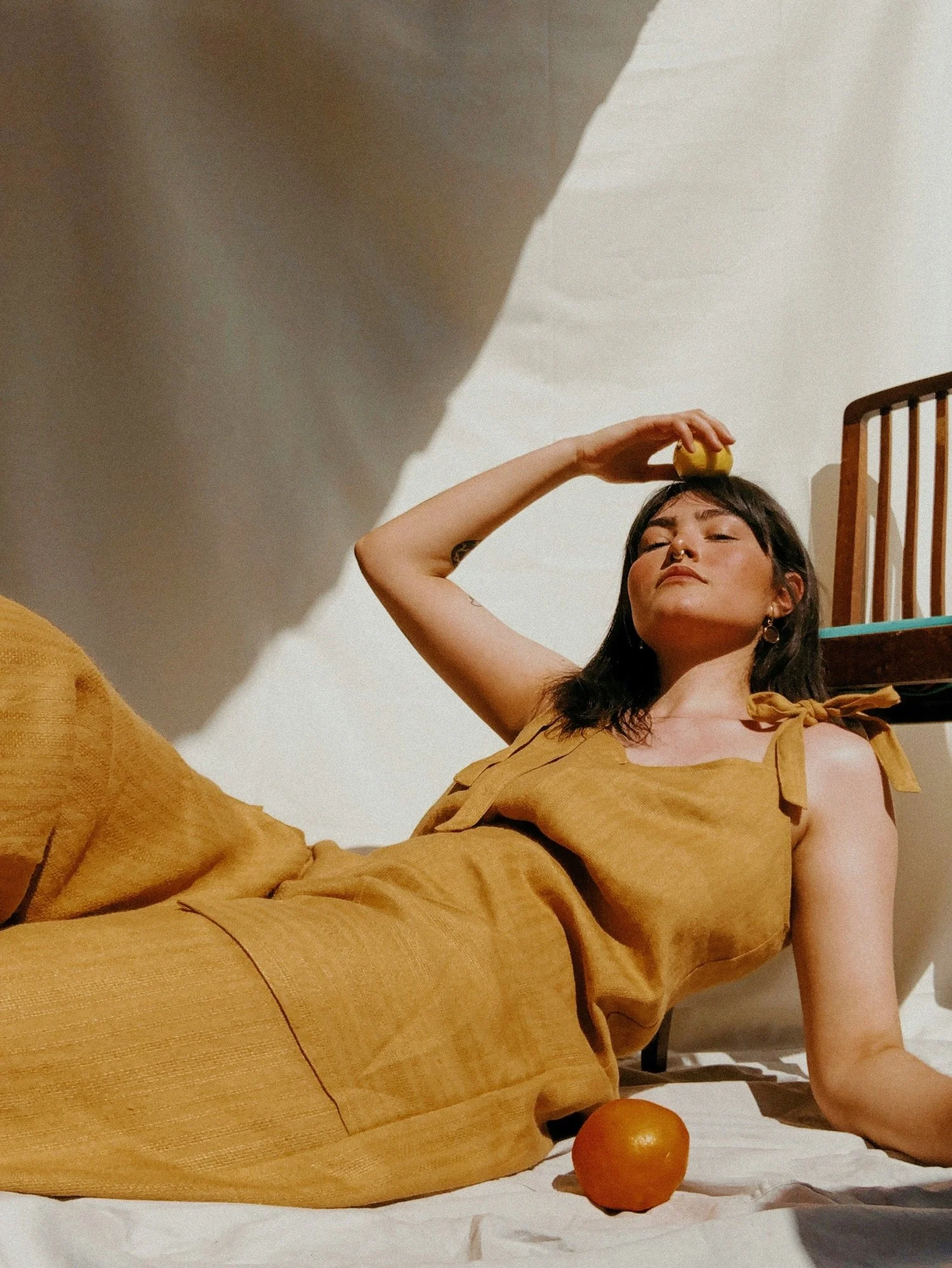 Young woman lying on her back on a white surface, holding a lemon above her forehead, with an orange on the surface near her and a cherry on her head. She is wearing a tan outfit and has a relaxed, confident expression.