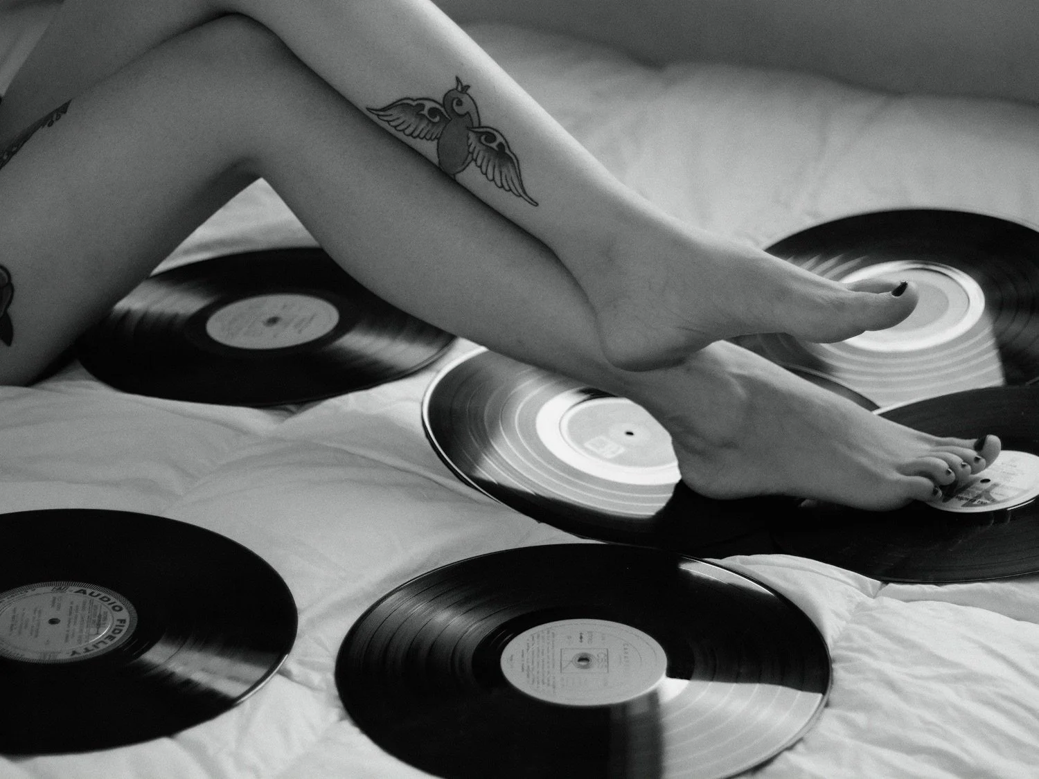 A person's arm with a tattoo of a bird and wings is resting on several vinyl records spread out on a surface, with one foot touching some of the records.
