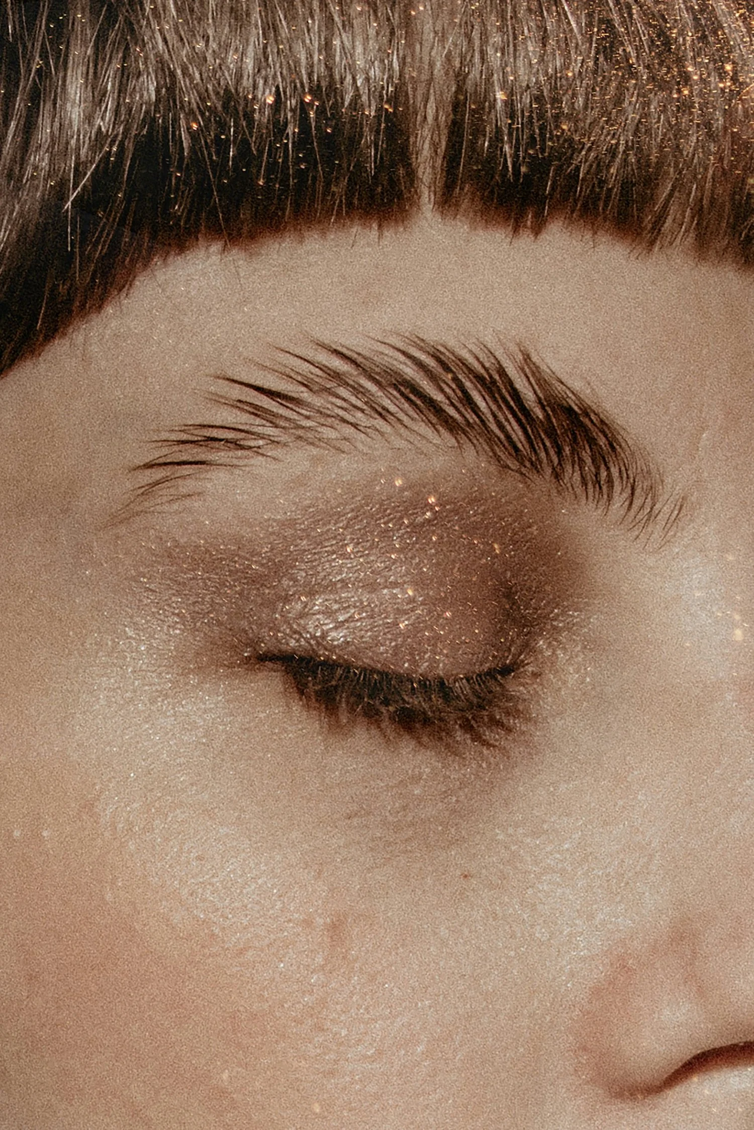 Close-up of a closed eye with shimmering gold eyeshadow and brown eyebrow, with short, straight brown hair in the background.