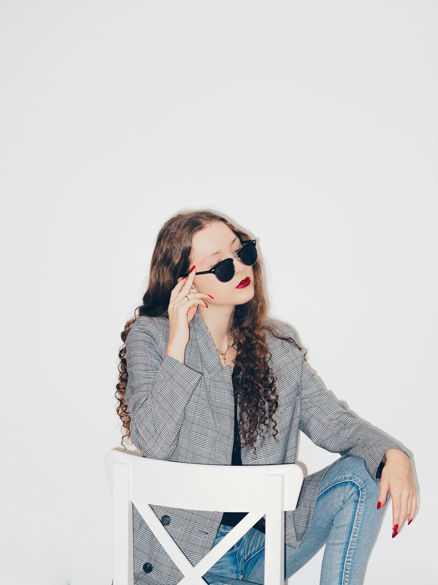 A young woman with long curly hair wears sunglasses, a plaid blazer, and blue jeans, sitting on a white chair with a plain white background.