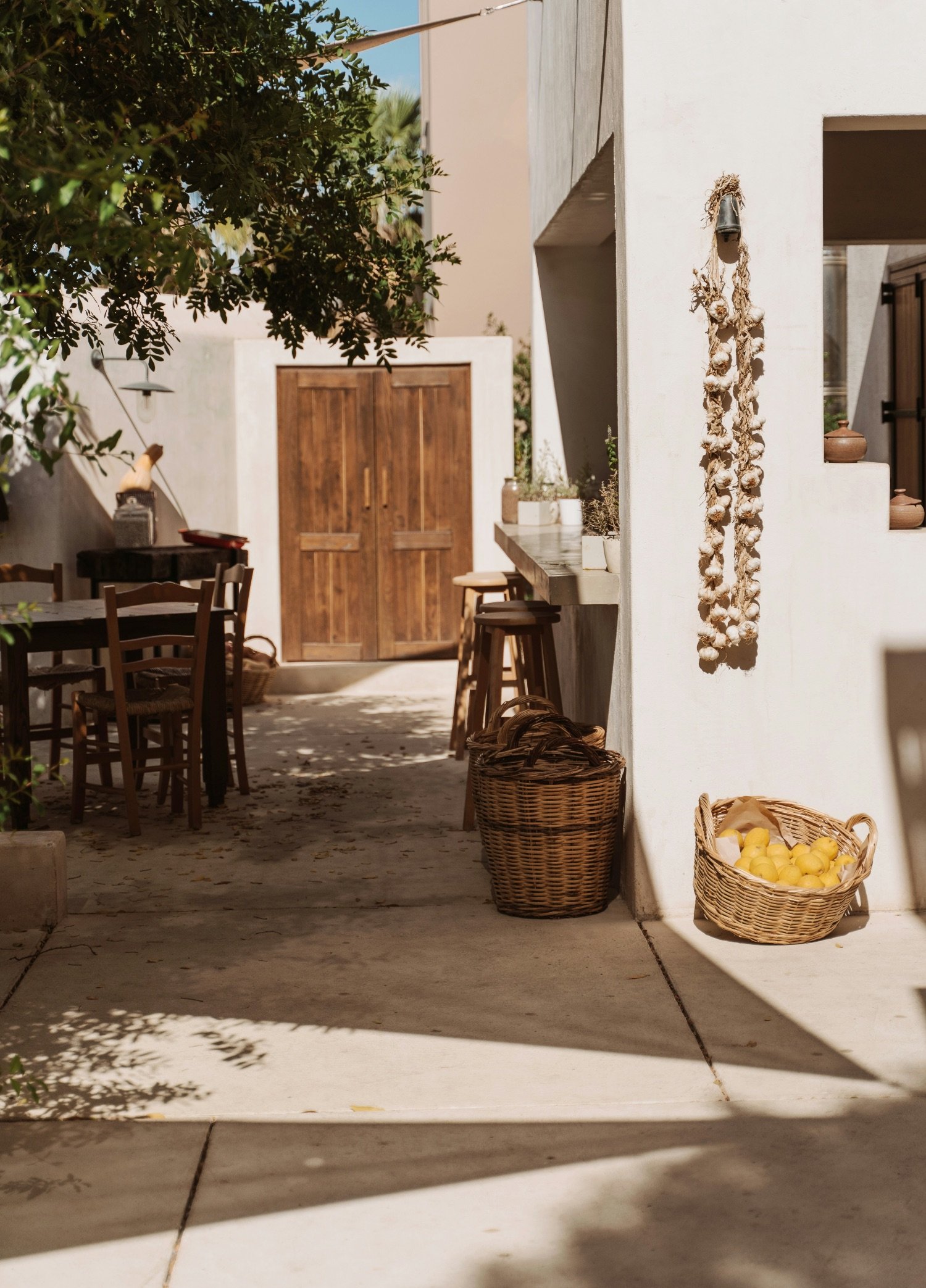 A cozy outdoor patio with wooden chairs, a table, baskets with yellow lemons, and hanging garlic braids, surrounded by a white stucco wall and lush greenery.