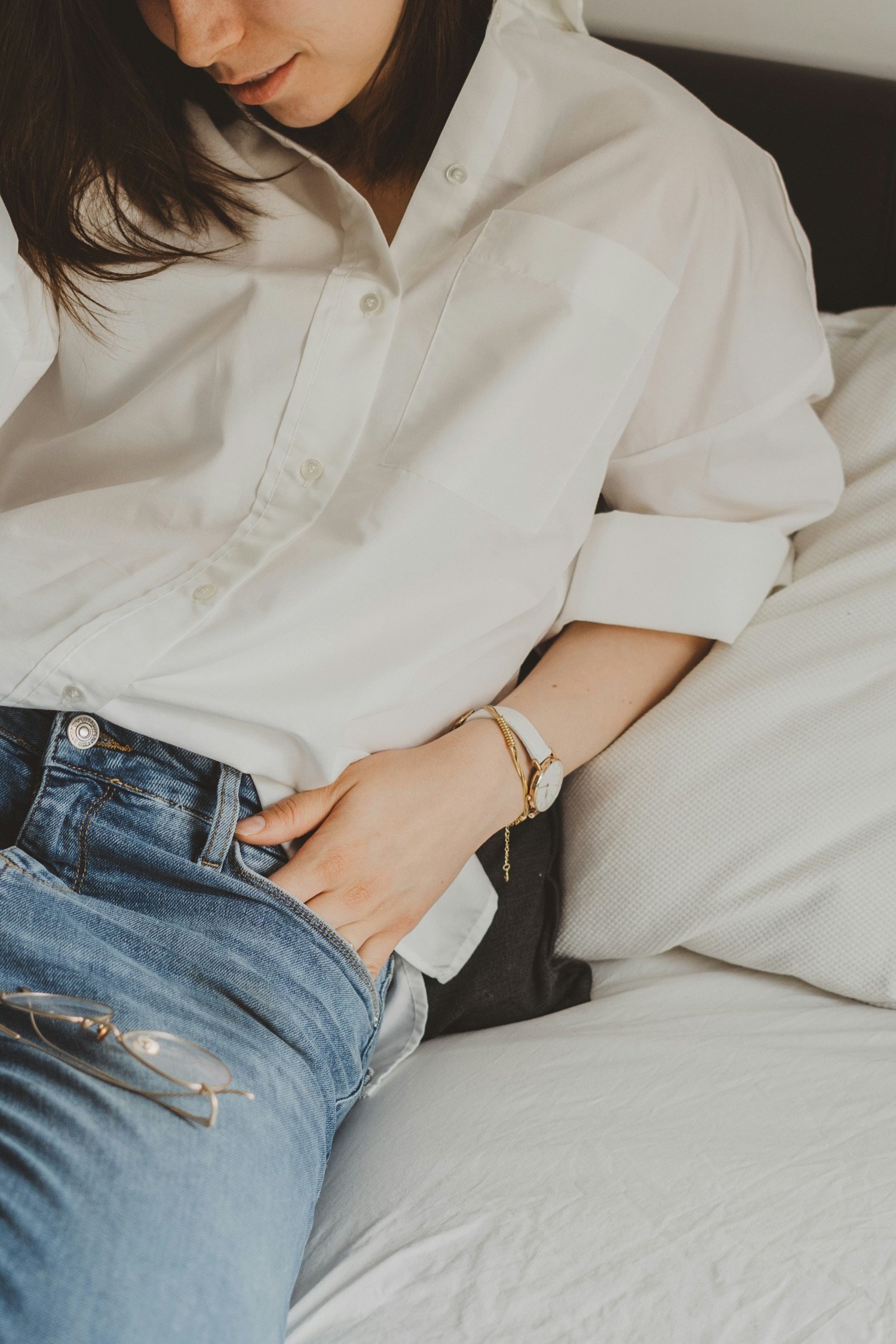 A person in a white oversized button-up shirt and blue jeans sitting on a bed, with sunglasses on their lap and a cuff watch on their wrist.