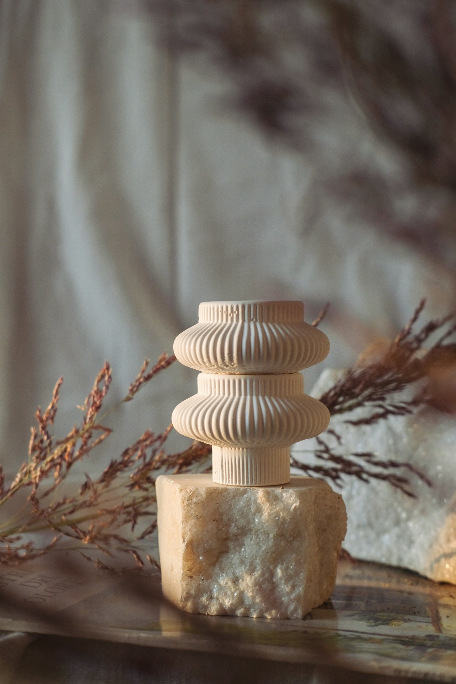 A beige sculptural object with layered, ribbed design sitting on a rough stone block, with dried plant stems in the background.