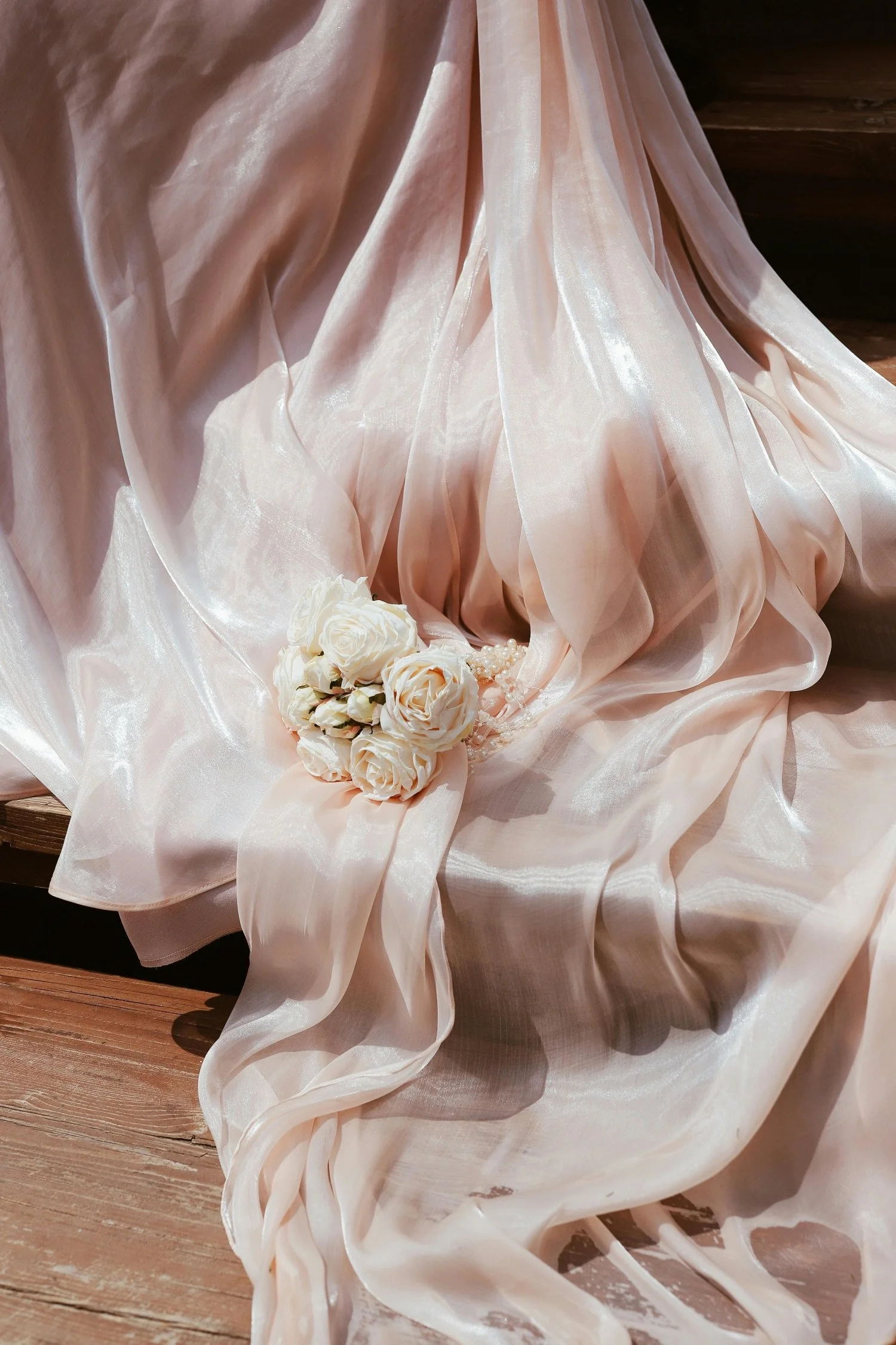 A soft pink satin bridal gown draped on a wooden surface with a small bouquet of white roses placed on it.