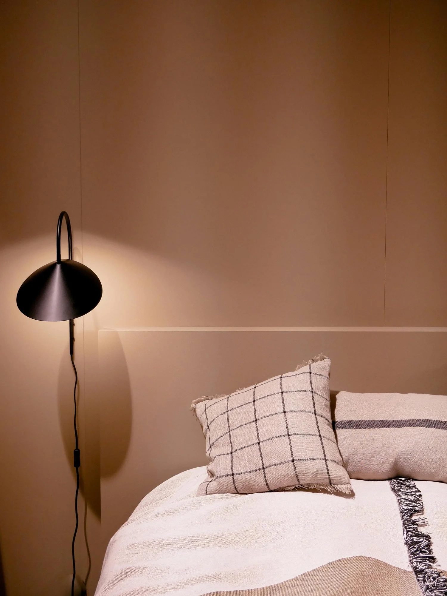 A bedroom corner with a black wall-mounted lamp turned on, beige wall, white bed with two decorative pillows, and a fringe blanket draped over the bed.