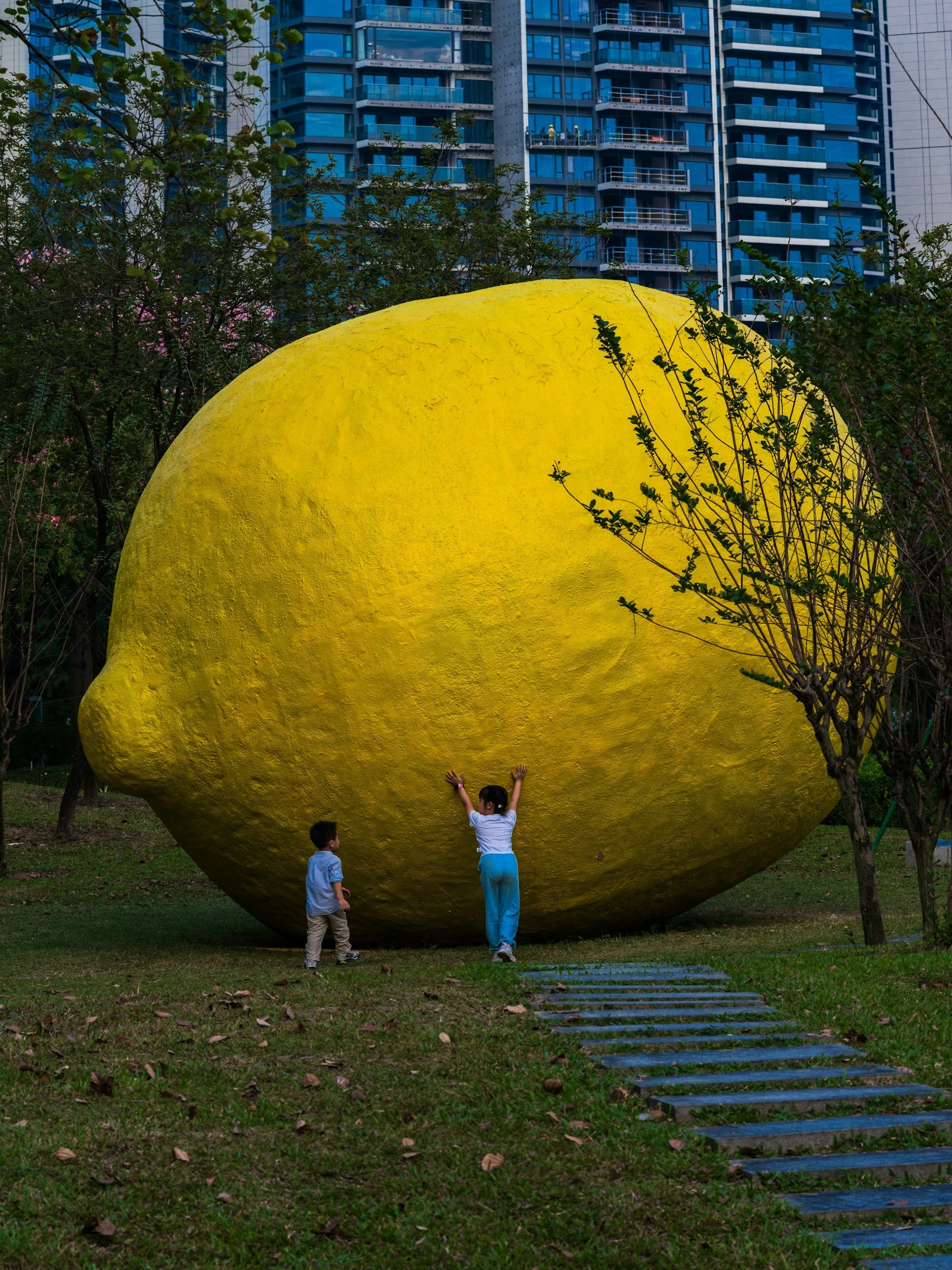 Children playing near a large, yellow, apple-shaped sculpture in an urban park with tall buildings in the background.