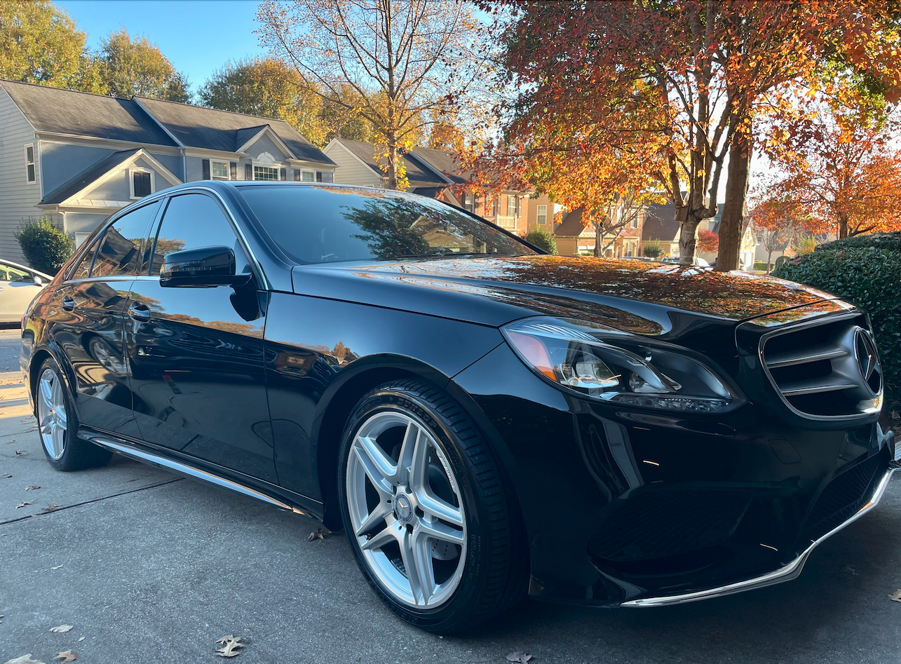 A shiny black Mercedes-Benz sedan parked in a driveway surrounded by trees with orange and yellow leaves, and suburban houses in the background on a sunny fall day.