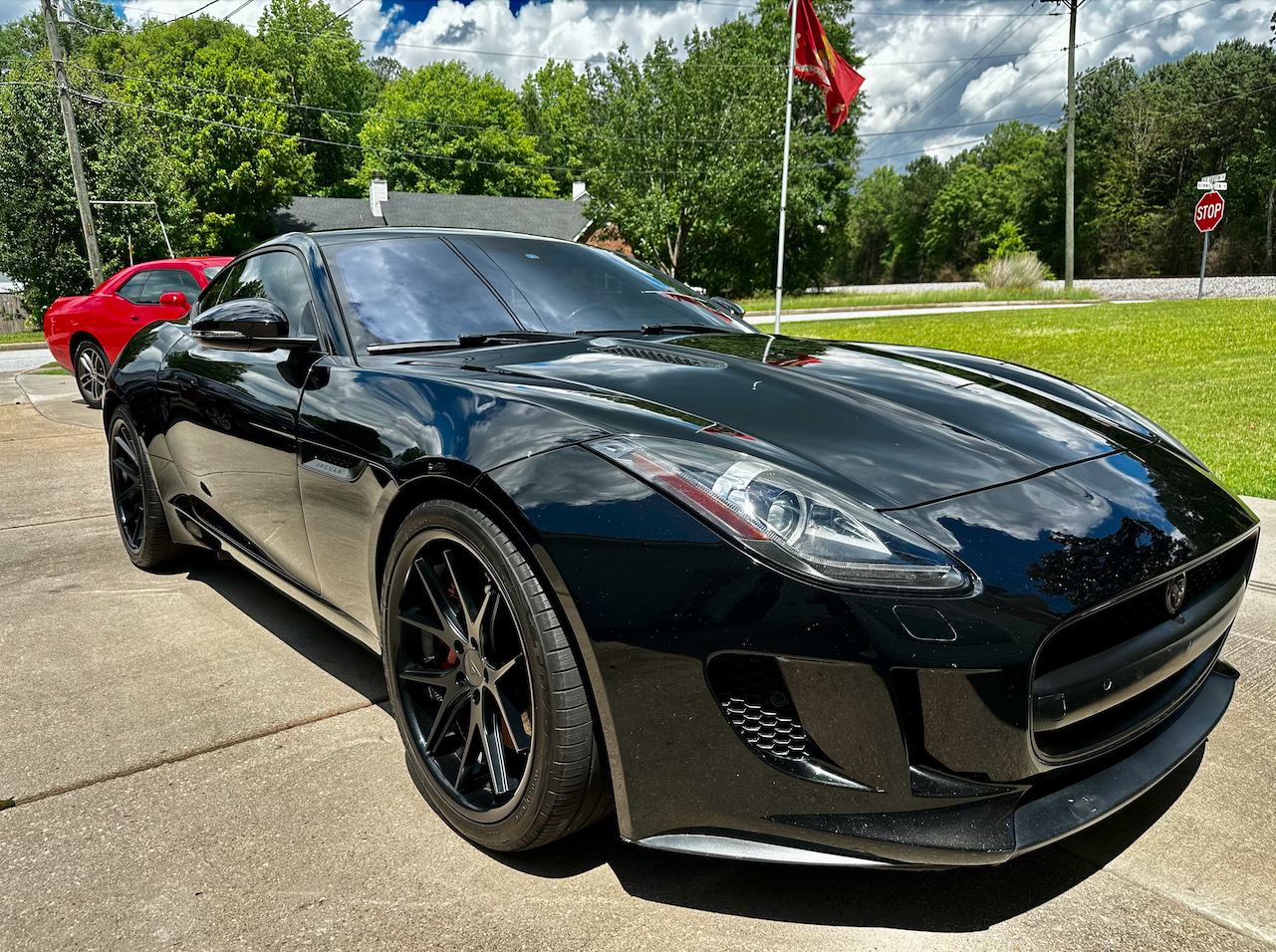 A black Jaguar F-Type sports car parked on a driveway with a red car behind it. The background features green trees, a house, flags, a stop sign, and a partly cloudy sky.