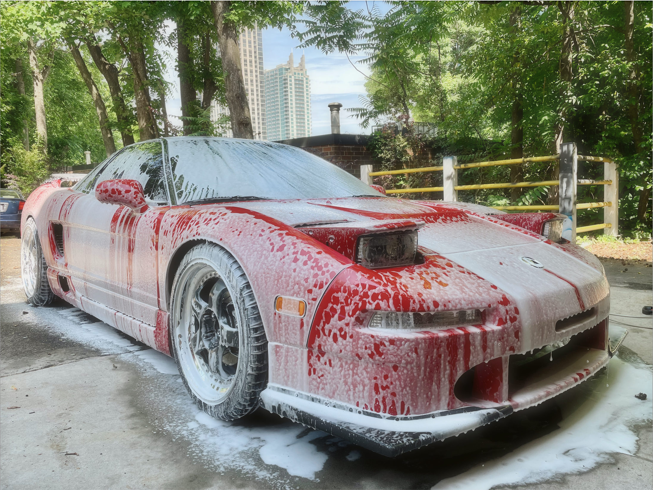 A red sports car is covered in soap suds and foam after being washed, with water and foam dripping off over a paved surface in an outdoor parking area surrounded by trees and tall buildings in the background.