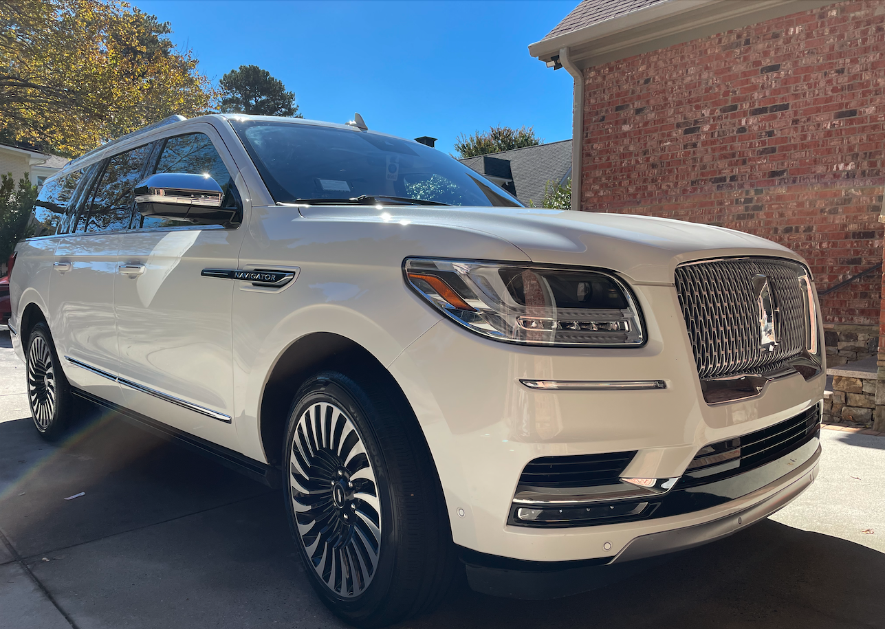 White Lincoln Navigator SUV parked in driveway next to brick house on a sunny day.