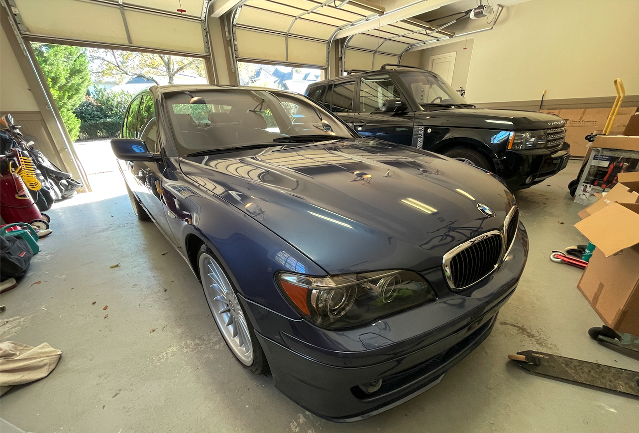 A dark-colored BMW convertible and a black Land Rover parked inside a garage, with garage doors open showing trees and sunlight outside, and various garage tools and boxes visible around.
