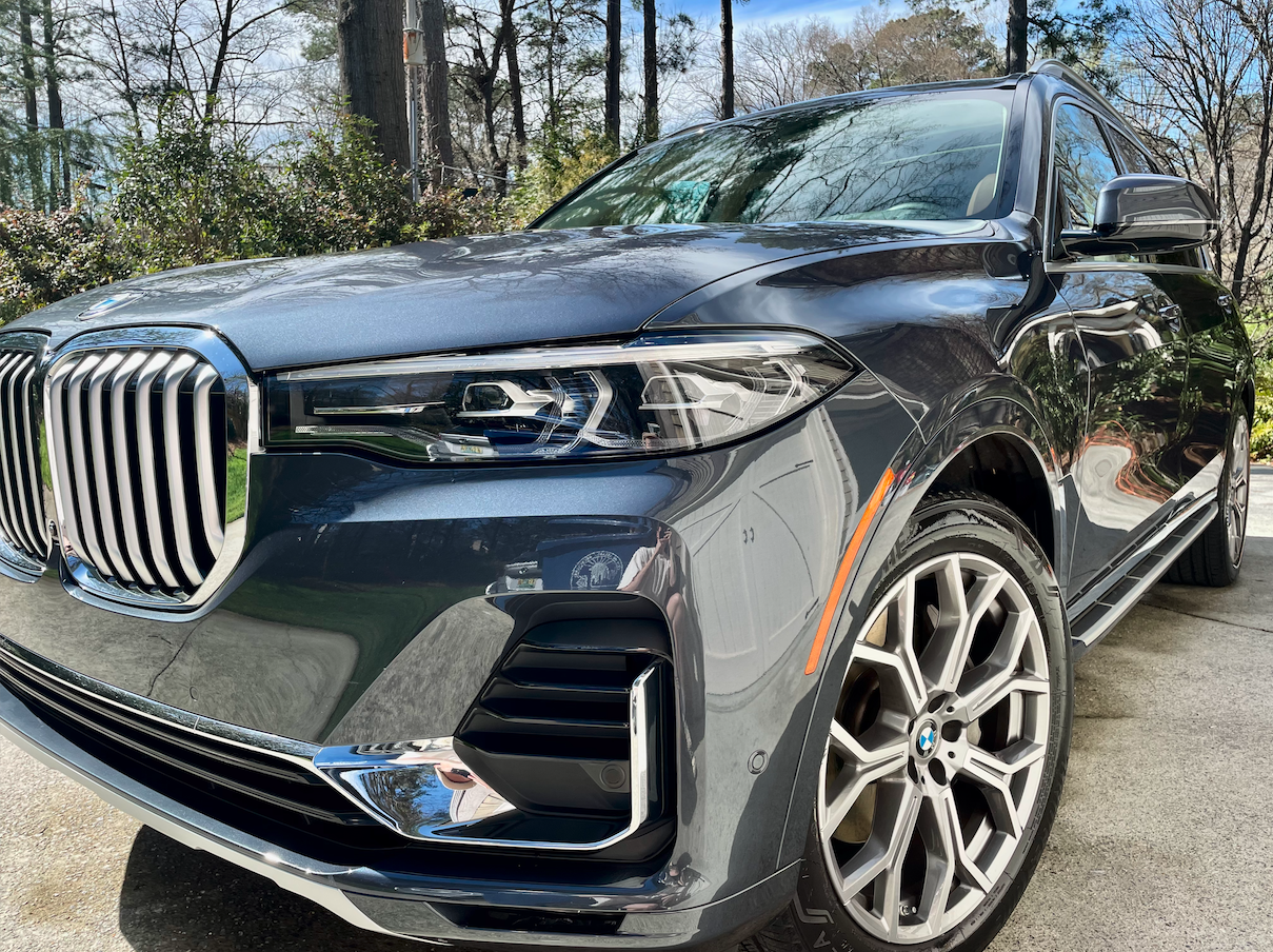 Close-up of a black BMW SUV parked outdoors, showing the front grille, headlight, and front wheel with trees and sky in the background.