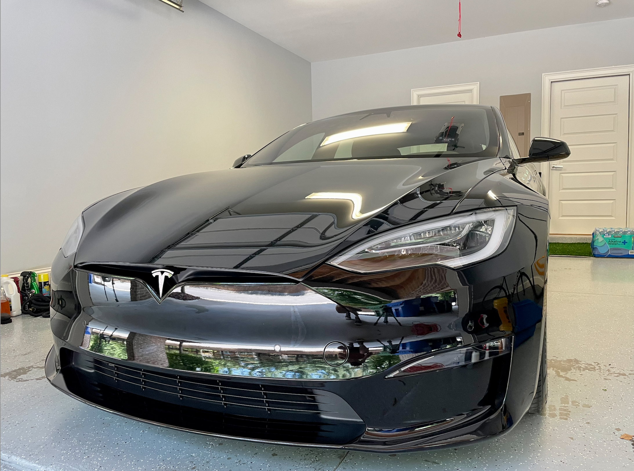 A black Tesla Model S parked in a garage, with garage walls and two doors visible in the background. The car's front is visible, showing the Tesla logo, headlights, and part of the windshield. The garage floor is speckled gray, with various tools and