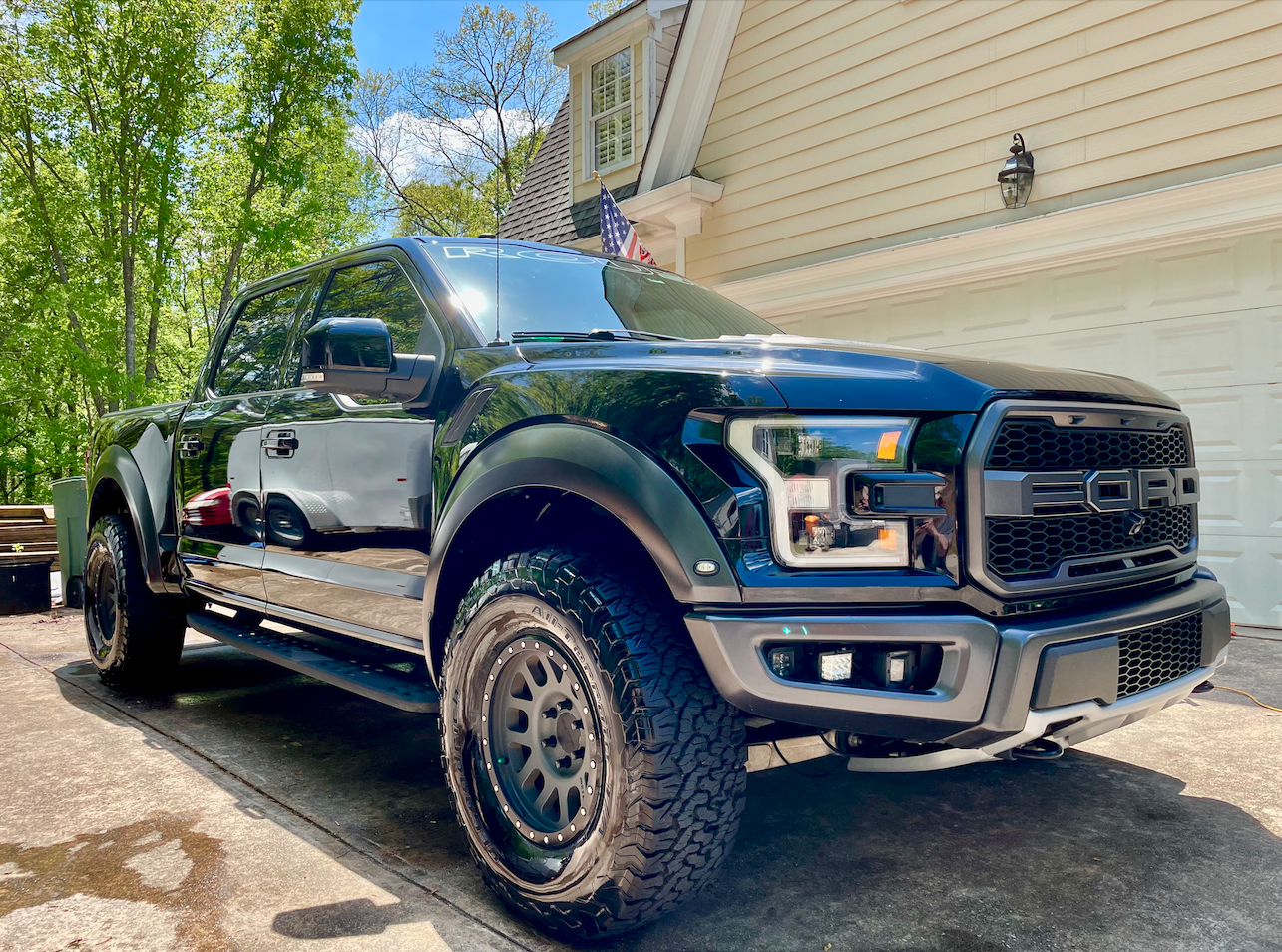 A black Ford F-150 Raptor pickup truck parked on a driveway in front of a beige house with a white garage door and an American flag, surrounded by green trees on a sunny day.