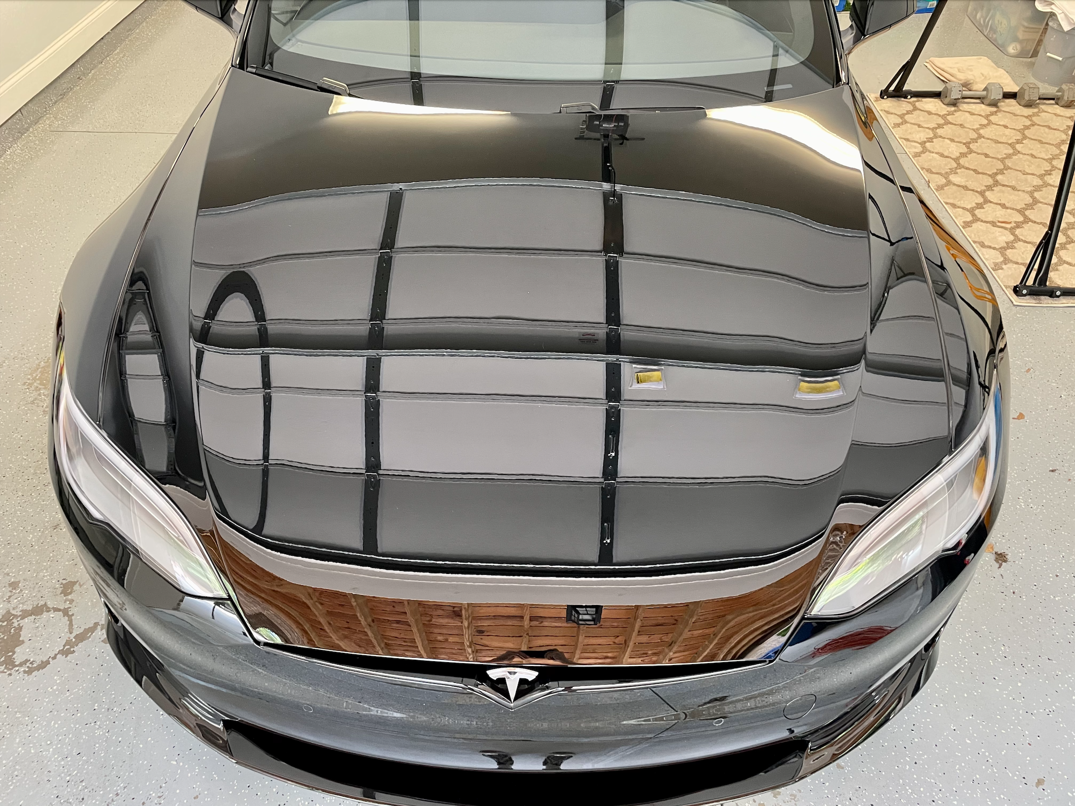 A black Tesla car parked in a garage with a two-toned roof reflecting the ceiling. The garage has a concrete floor, a shelf with items, and gym equipment. The car's hood shows a sleek, shiny surface with reflections of the garage interior.
