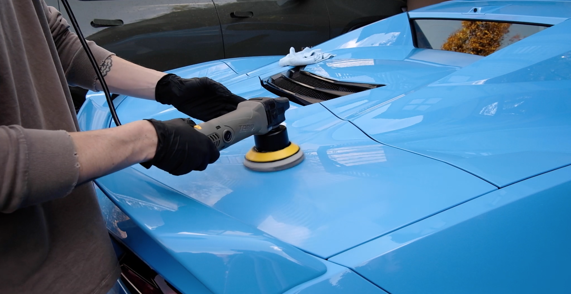 Person polishing the hood of a light blue sports car with a power buffer, wearing black gloves and a brown shirt.
