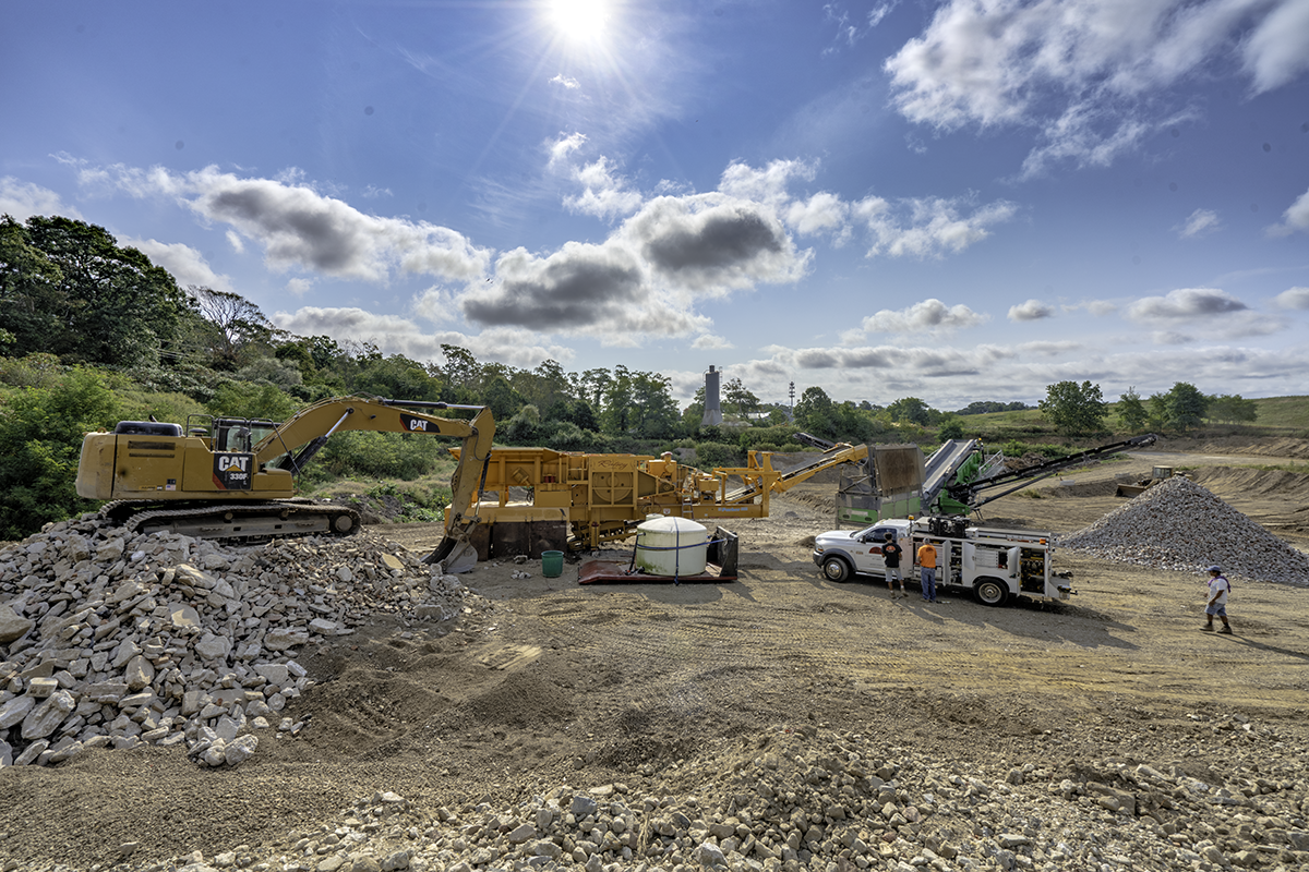 Construction site with excavator, conveyor belt, and truck working on gravel and rocks under a partly cloudy sky.