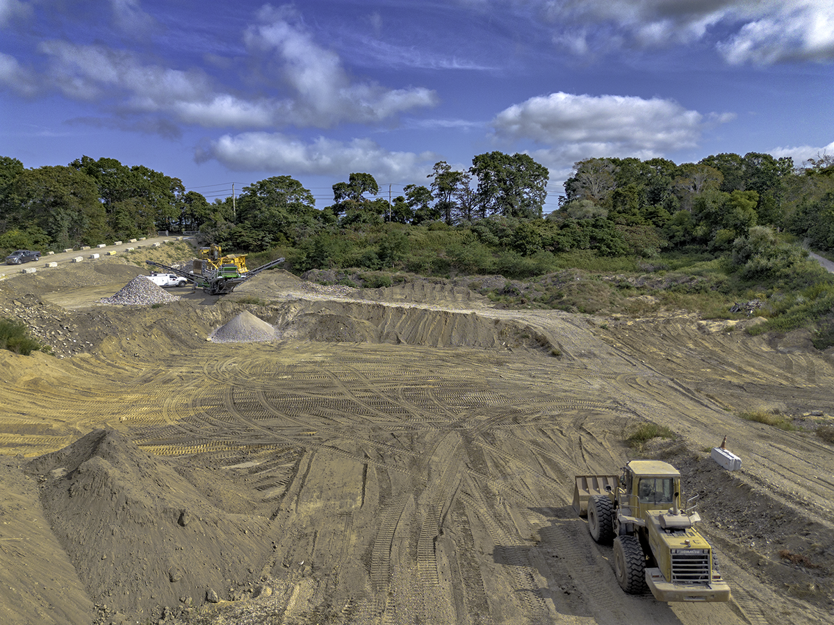 Construction site with a bulldozer in the foreground, piles of dirt and gravel, and trees with a partly cloudy sky in the background.