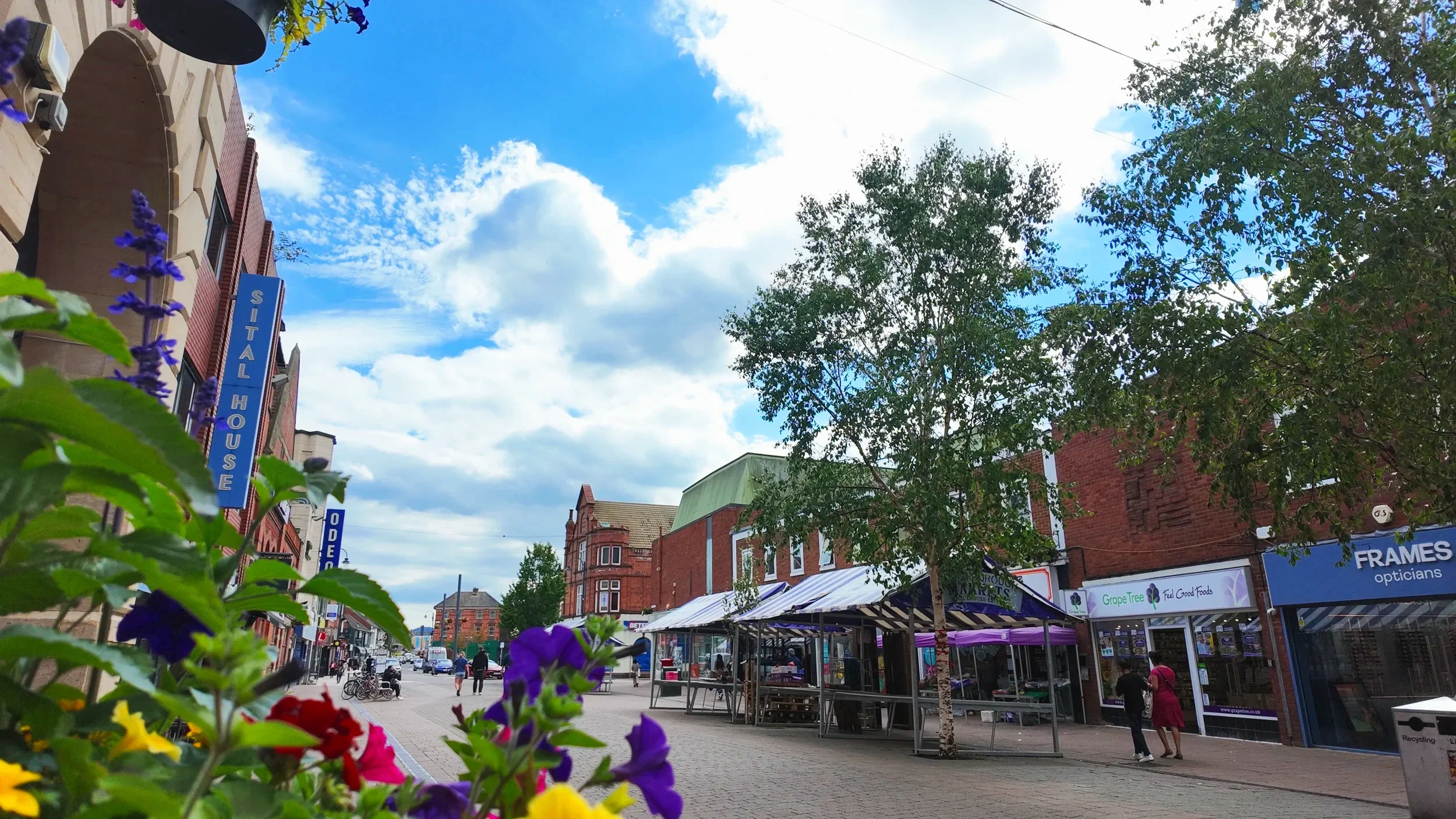 A lively city street scene with storefronts, outdoor market stalls, and colorful flowers in the foreground. The sky is partly cloudy with blue patches, and there are trees and buildings lining the street.