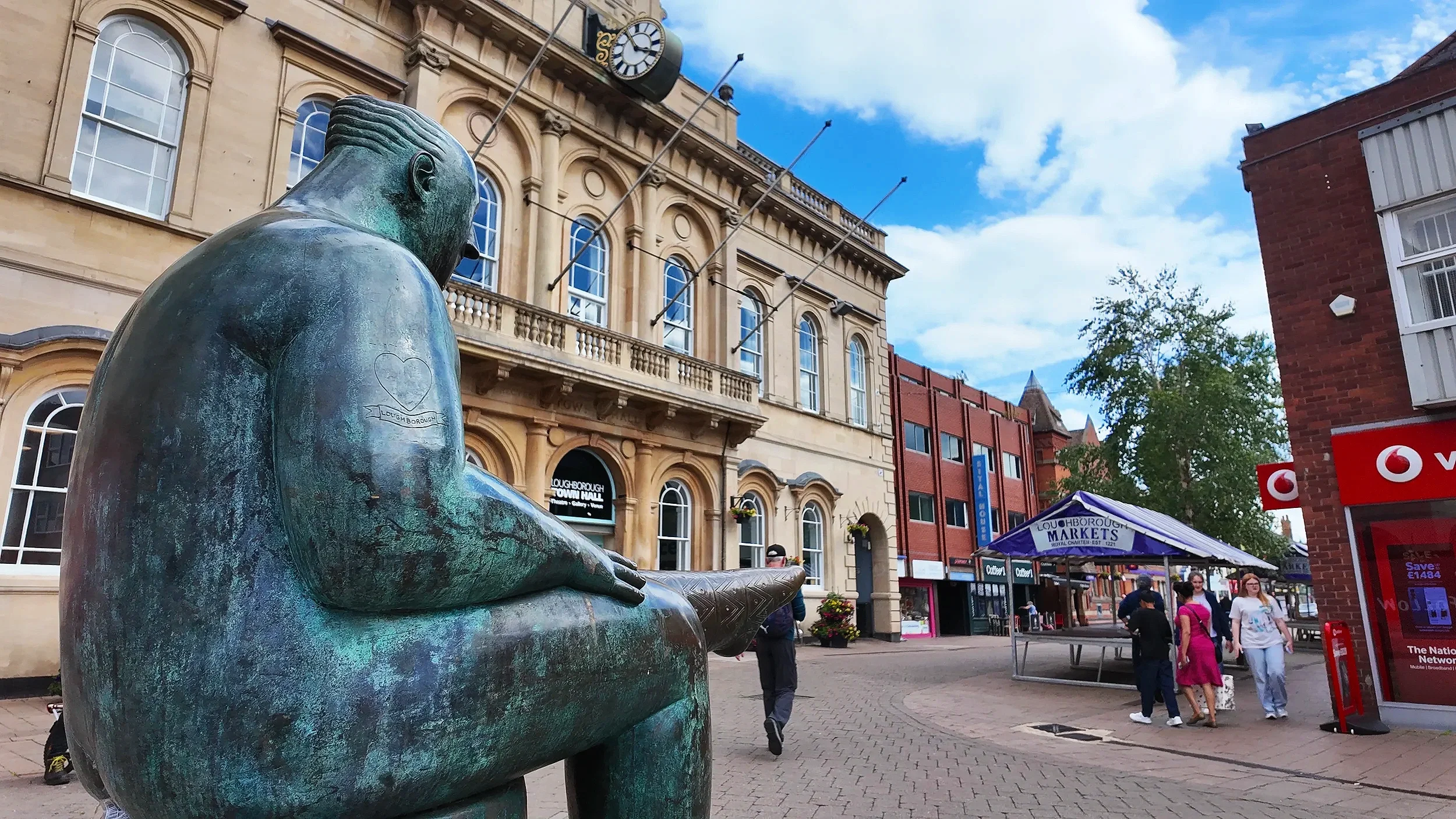Bronze sculpture of a boy sitting with his arms crossed, located in a busy town square with historic buildings, shops, and people walking and standing around.