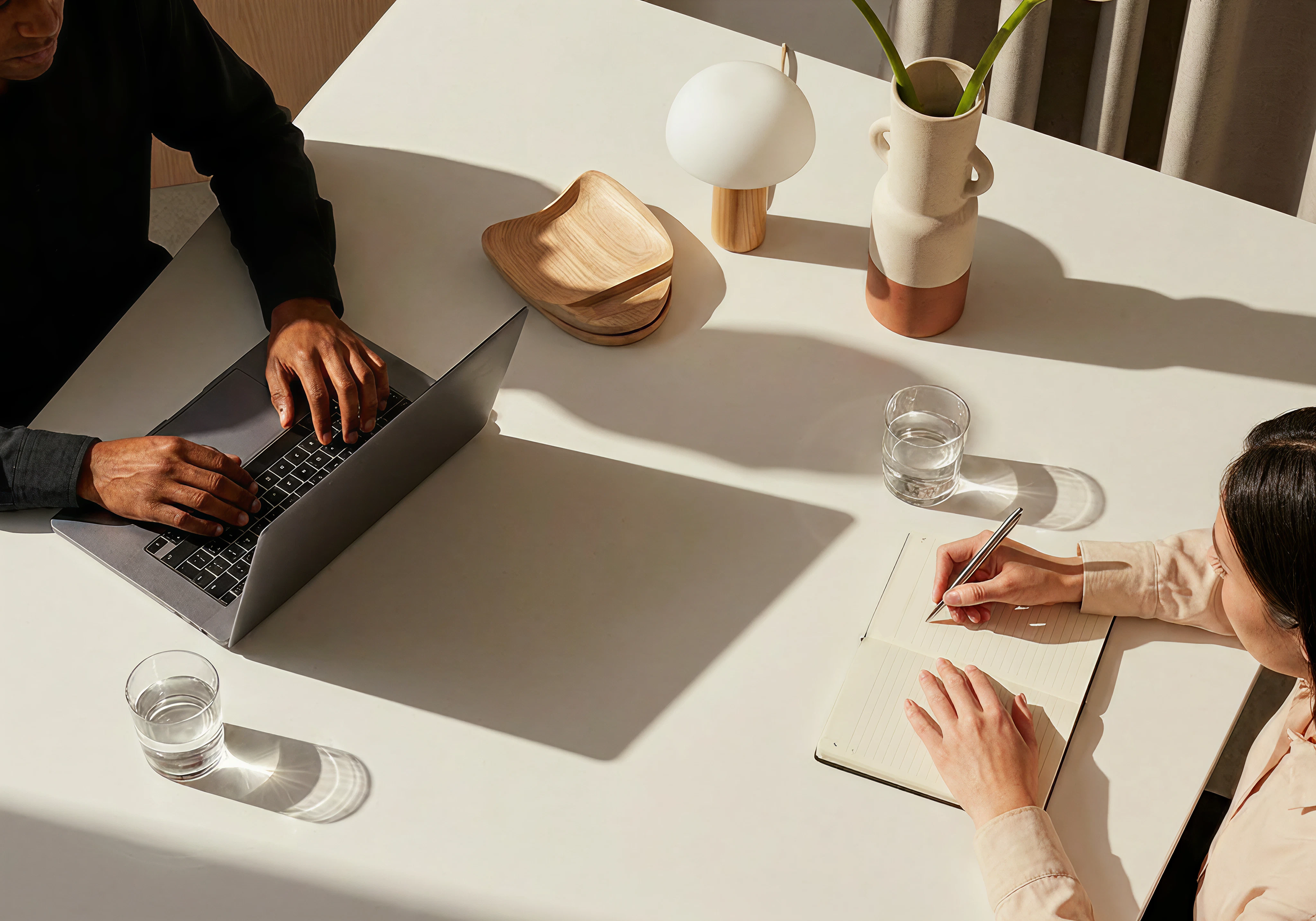 A person working on a laptop at a white table, with a notebook and pen, and a child writing. On the table are three decorative vases, one with a plant, and two glasses of water.
