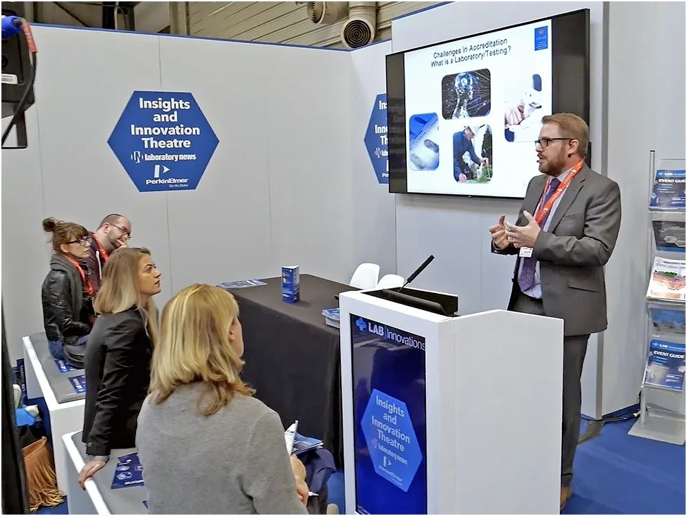 A man in a gray suit giving a presentation at a conference booth, with a large screen displaying slides titled 'Challenges in Accreditation What is Laboratory Testing?'. Six people are sitting and listening, some taking notes, in front of the presenter.