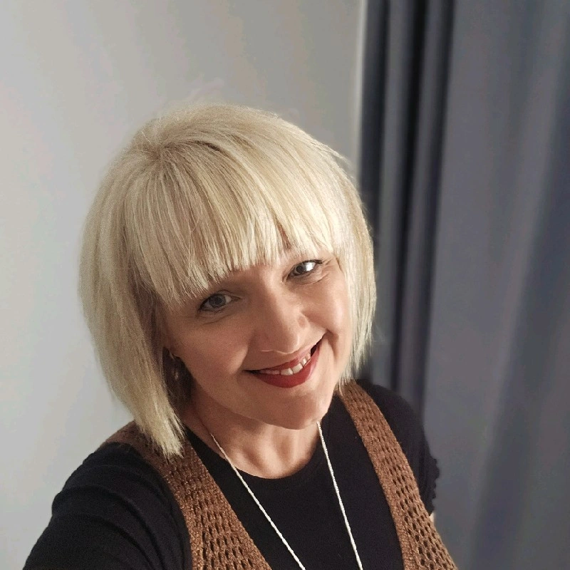 A smiling woman with short, blonde hair wearing a black top and brown vest, standing indoors near a door.