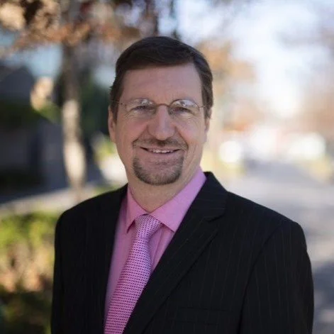 A man in a black suit with a pink shirt and tie, standing outdoors in a sunny area with trees in the background.