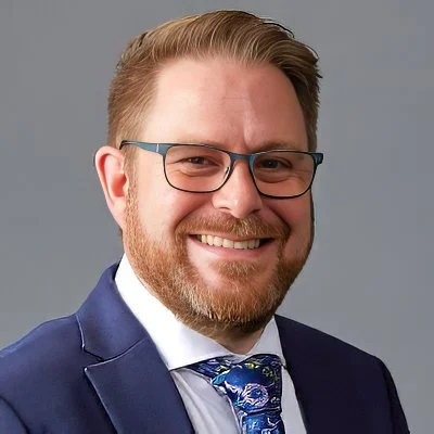 Headshot of a smiling man with glasses, a beard, wearing a navy suit, white shirt, and a patterned blue tie against a gray background.