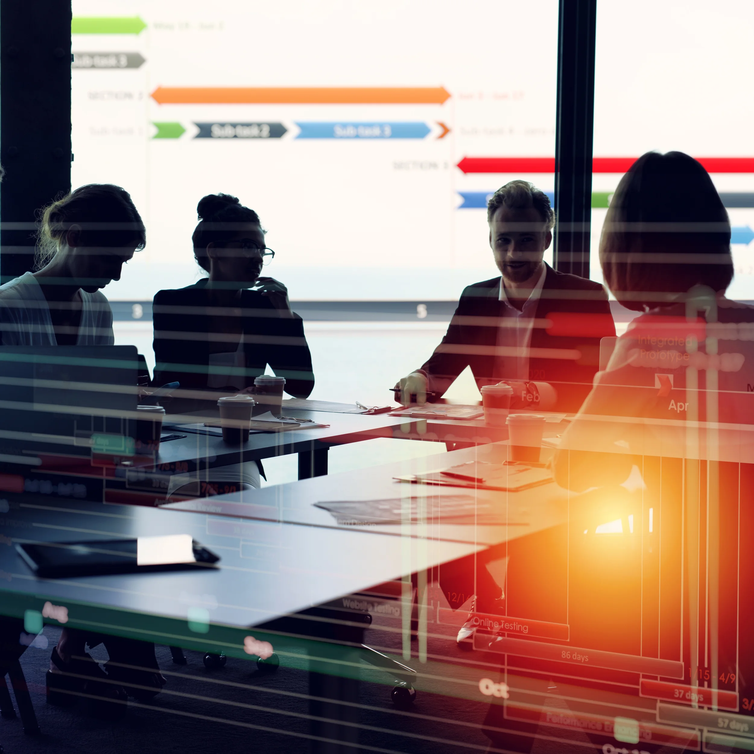 A group of five people sitting and standing around a table having a discussion in a bright room, with a large colorful timeline or schedule chart projected on the window behind them, displaying months and dates.