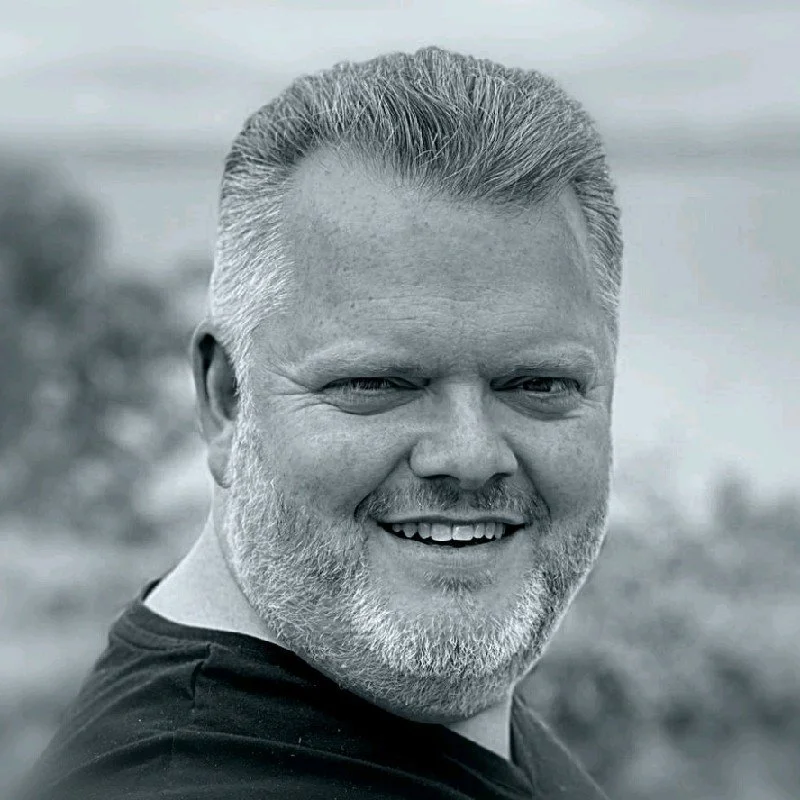 A close-up black and white portrait of a smiling middle-aged man with short, neatly combed hair and a beard.