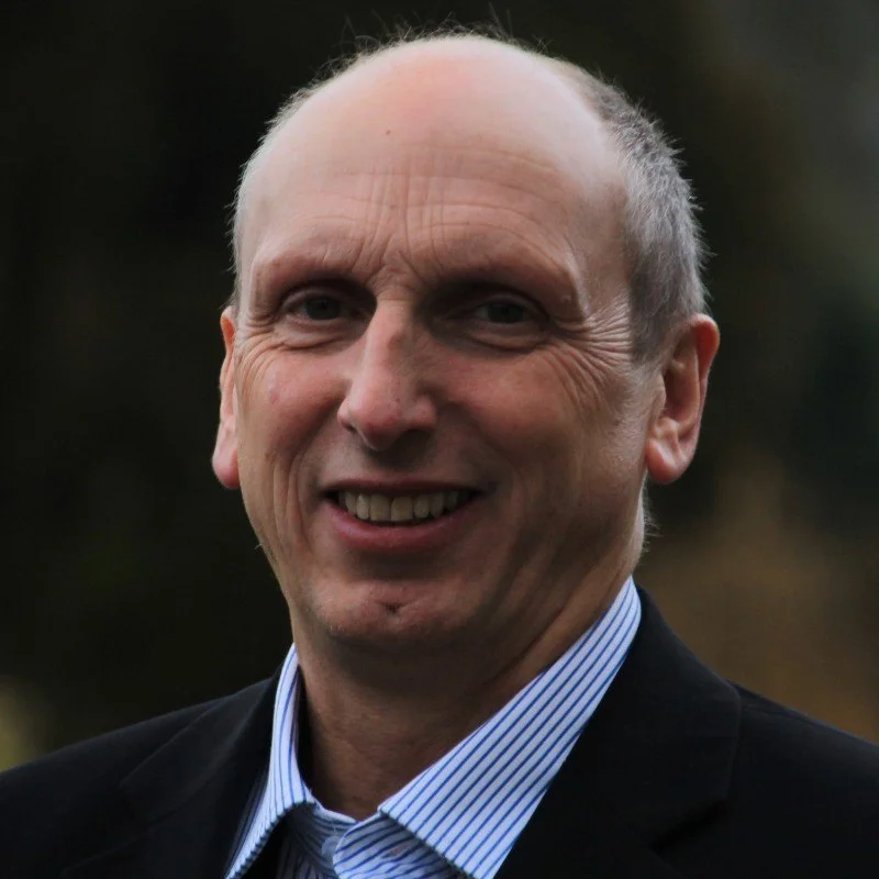 A middle-aged man with a bald head and a friendly smile, wearing a dark blazer and a striped dress shirt, outdoors against a blurred natural background.