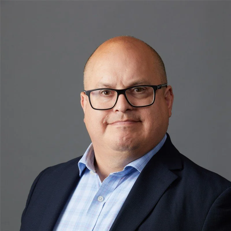 Professional portrait of a bald man with glasses wearing a suit jacket over a light blue dress shirt, gray background.