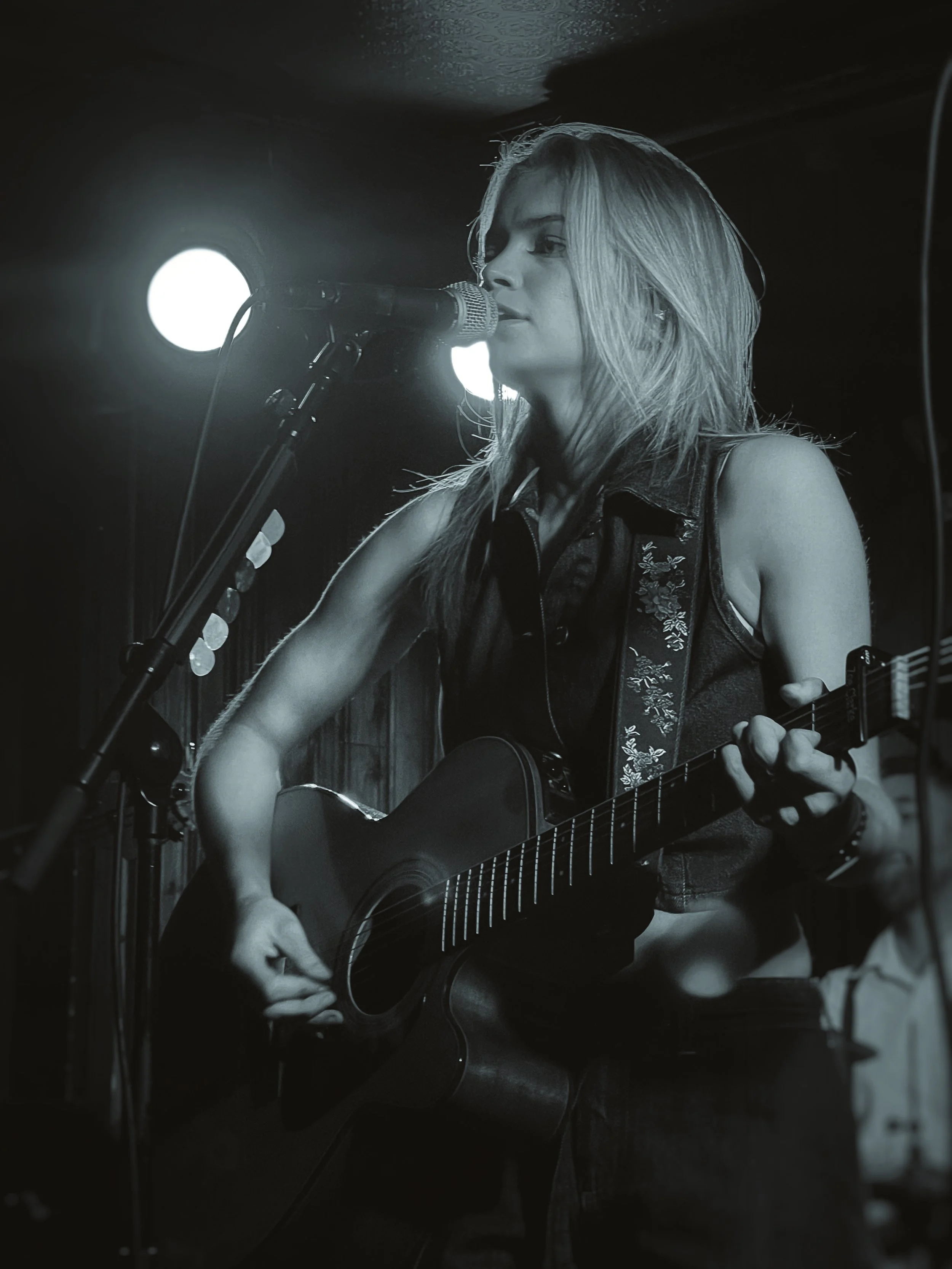 A woman with long blonde hair playing an acoustic guitar and singing into a microphone in a dimly lit room, black and white photo.