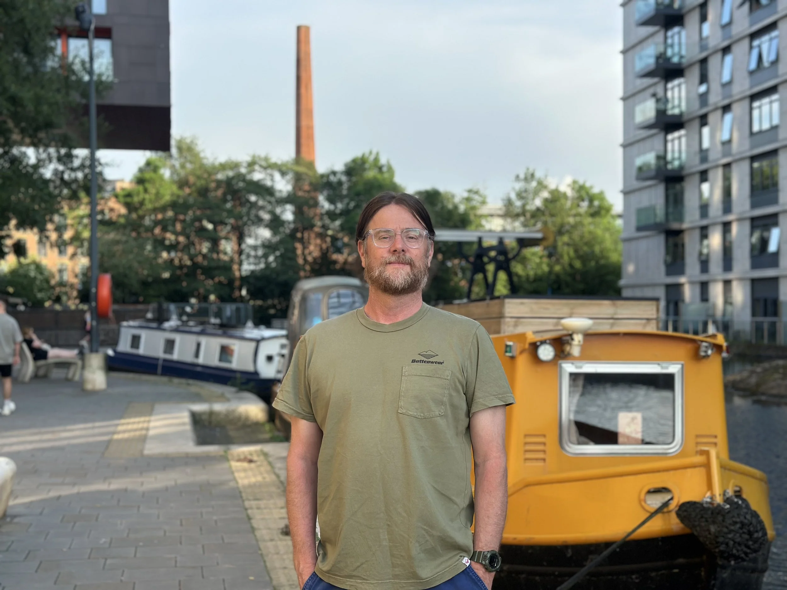 A man with glasses and a beard wearing a green T-shirt standing outdoors near a yellow machine, with modern buildings, trees, and a river in the background.