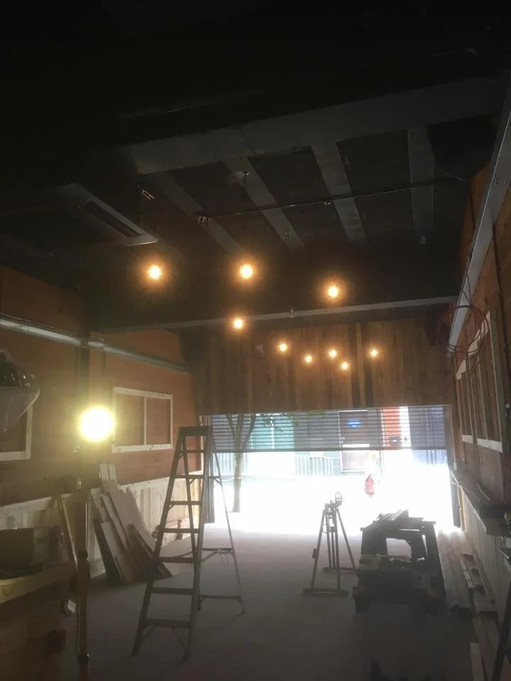 Interior of a building under construction or renovation, with ladders, tools, and lighting fixtures installed, illuminated by natural light coming through an open entrance.