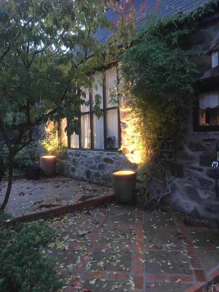 A stone exterior wall with large windows, surrounded by greenery and outdoor lighting, on a brick patio with fallen leaves.
