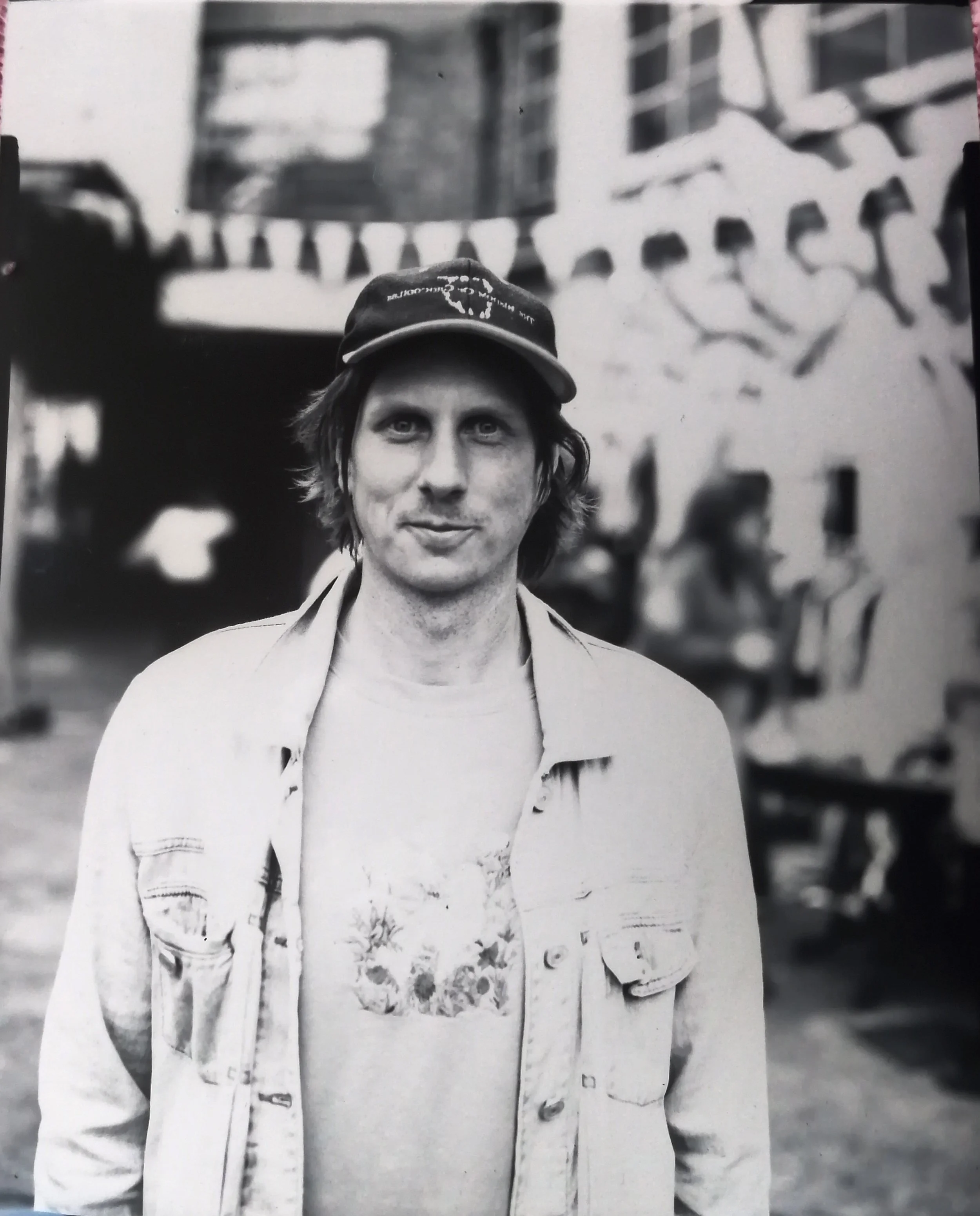 Black and white photo of Hitch Seed founder Thom Hoffman with medium-length hair, wearing a baseball cap, a light-colored jacket, and graphic T-shirt, standing in front of a blurred background.