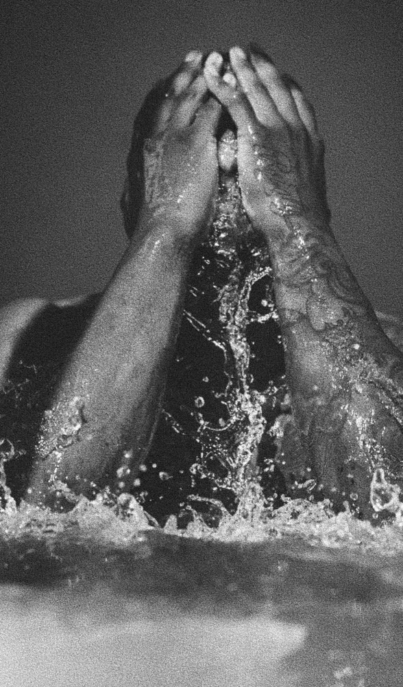 Person covering their face with both hands in water, water splashing around, black and white photo.
