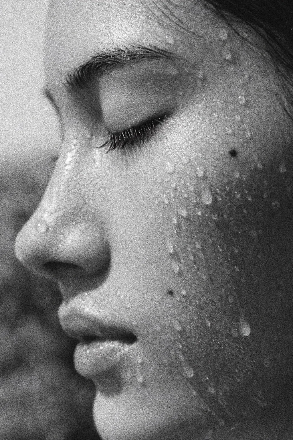 Close-up black and white photograph of a woman's face in profile with her eyes closed, covered in water droplets.