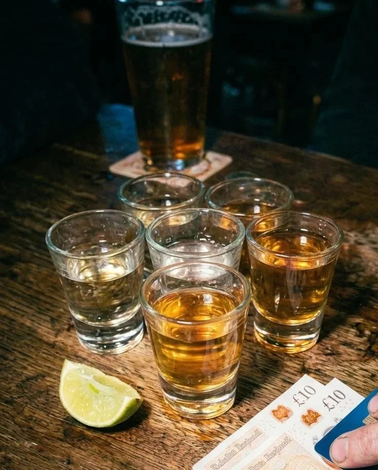 A round of shots on a table, with a pint of beer, £10 notes and a wedge of lime on the table