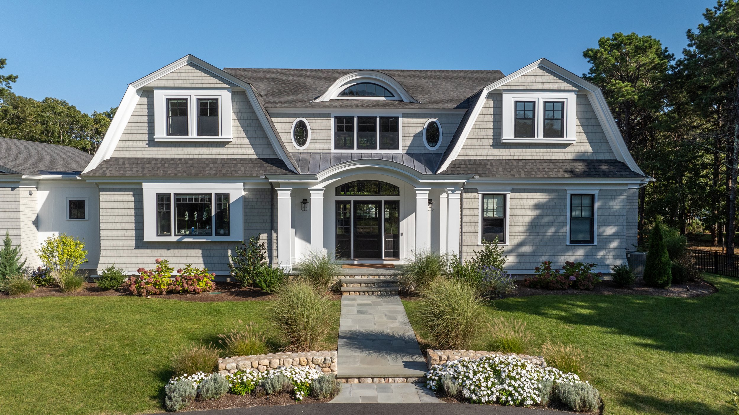 Front exterior of new construction shingle-style home on Monomoscoy Island, Mashpee, featuring a gambrel roof with arched eyebrow dormer, oval windows, columned entry portico, and bluestone walkway through hydrangeas.  James Phillip Golden Architect